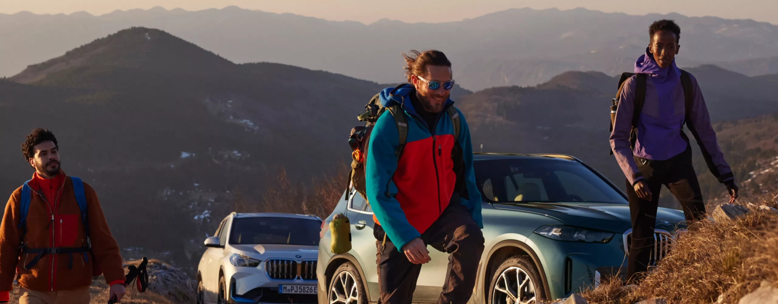 3 persons are climbing a mountain, there are two BMW vehicles parked in the background in front of a mountain vista