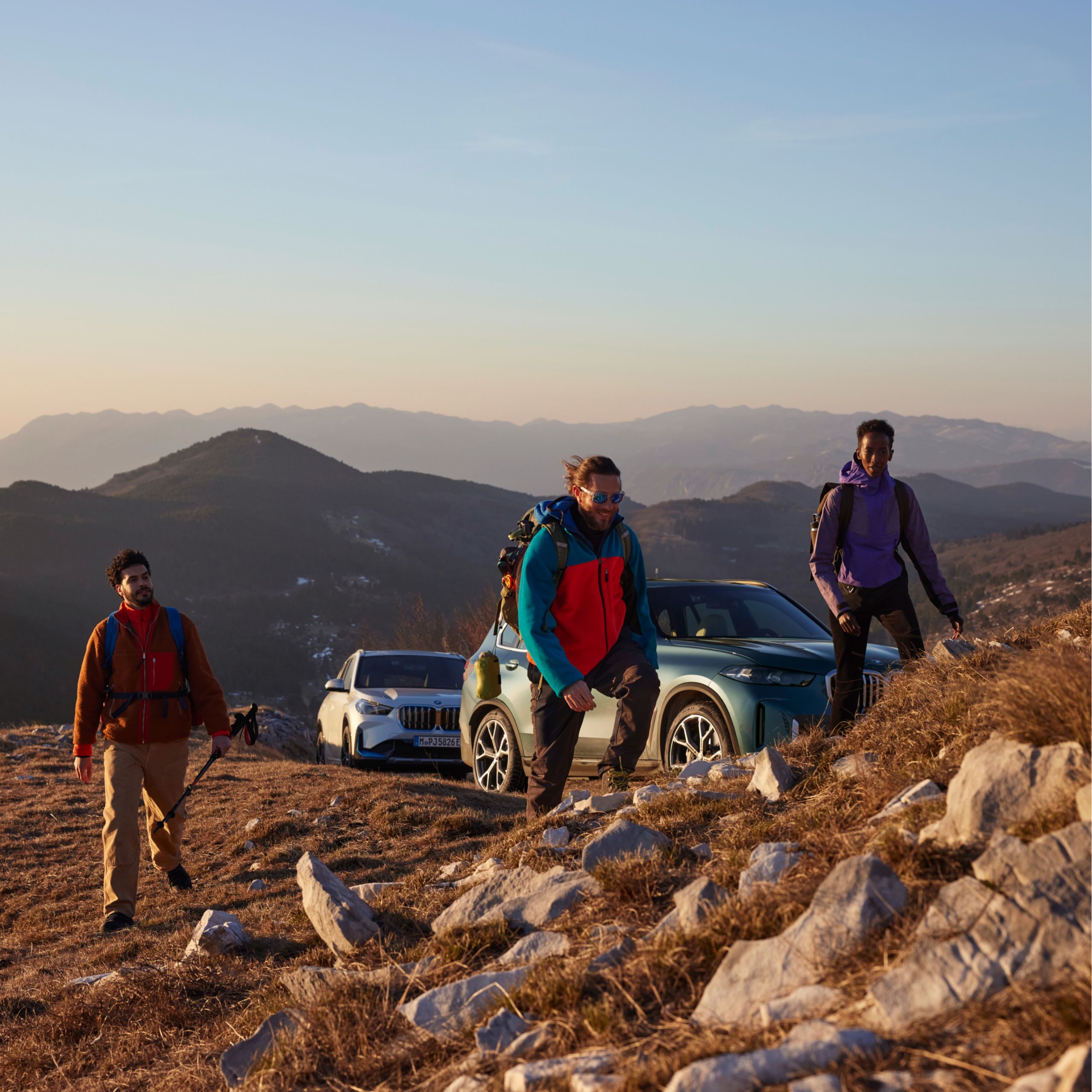 3 persons are climbing a mountain, there are two BMW vehicles parked in the background in front of a mountain vista