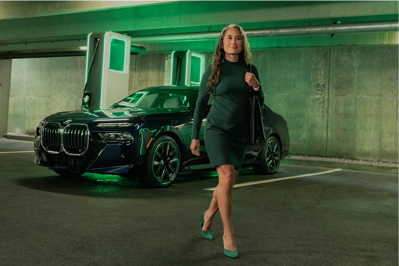 Inside a parking garage, a woman walks away from her BMW i4 as it charges at a public charging station.