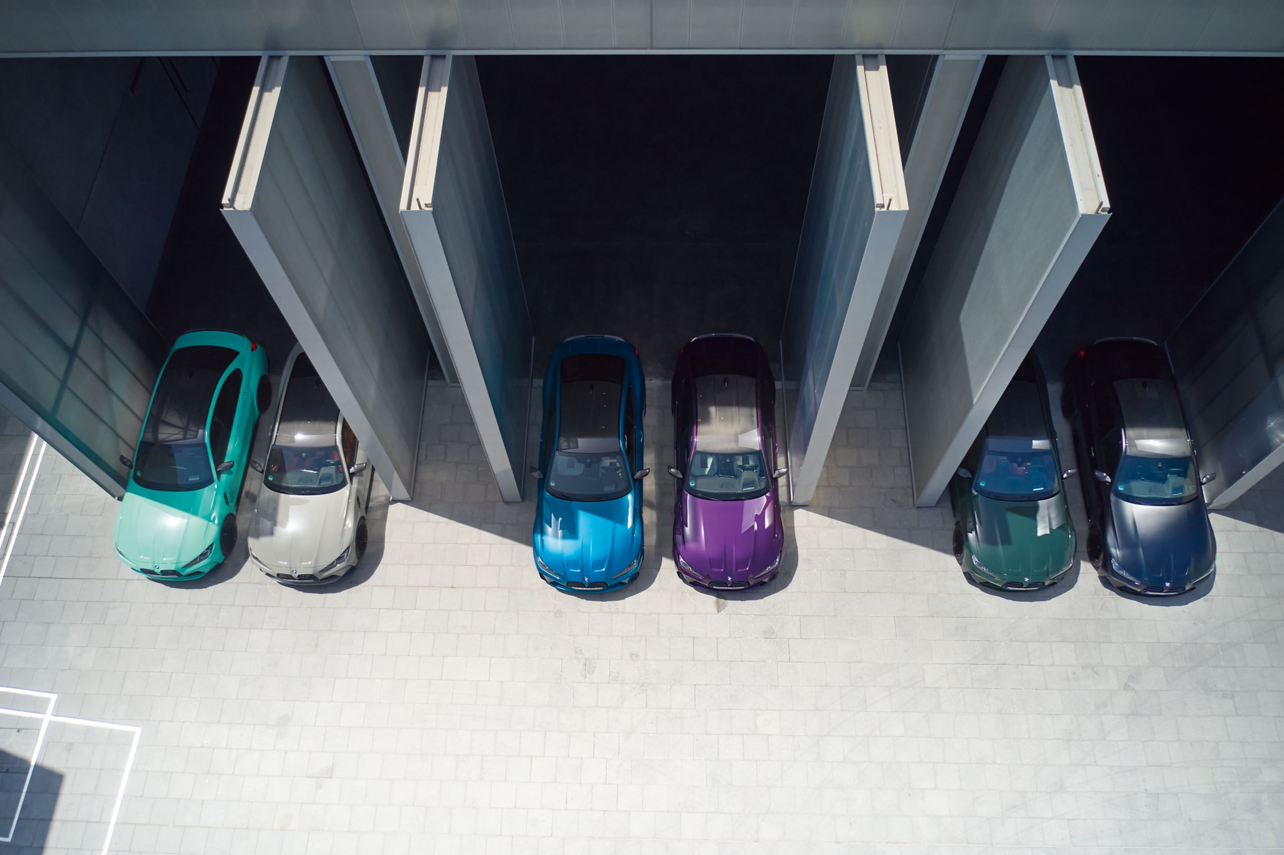 Overhead view of five brightly colored cars—turquoise, silver, blue, purple, and green—parked under a modern structure, casting sharp shadows.