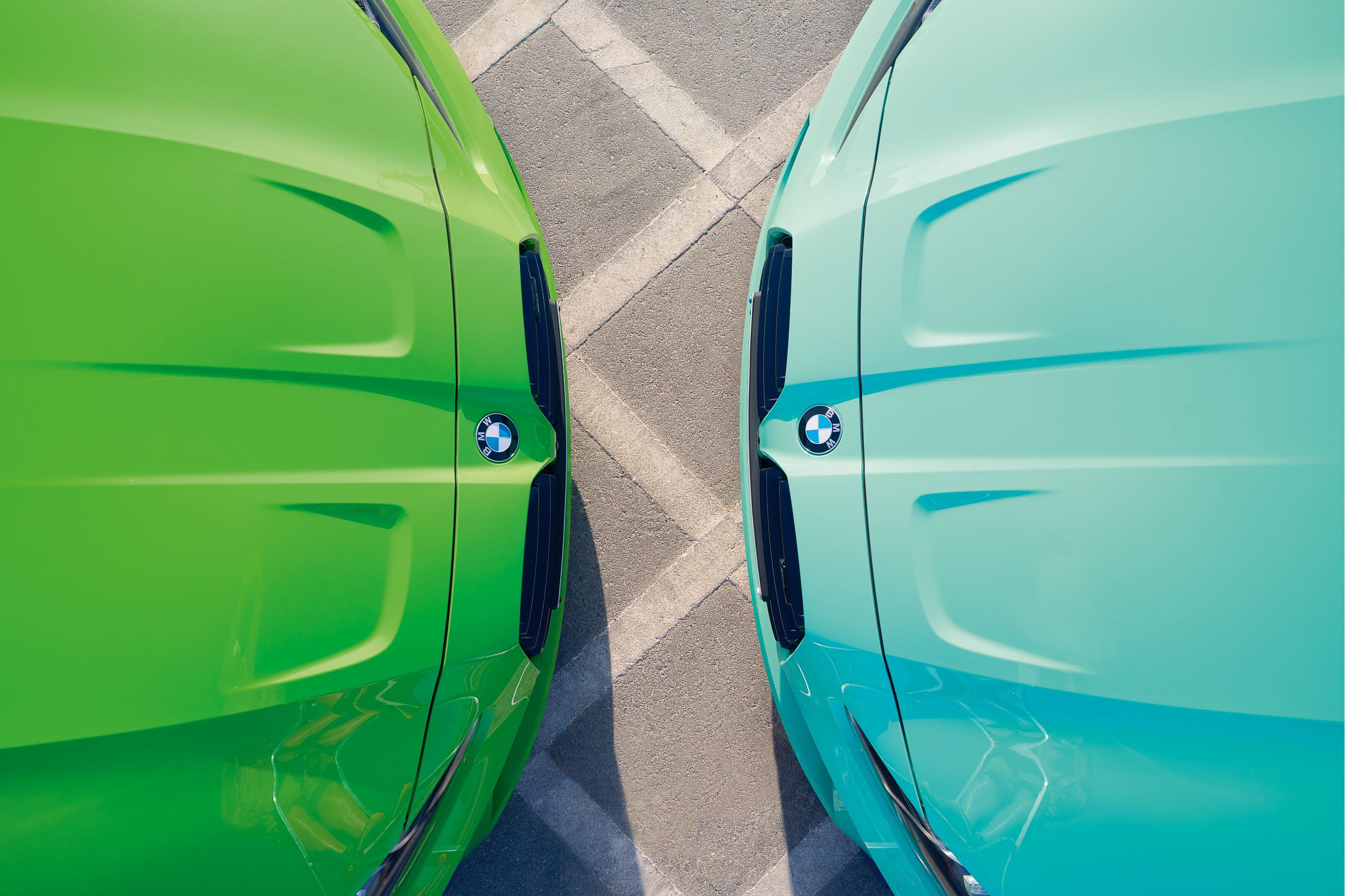 Overhead view of two BMW car hoods in vibrant green and teal, parked closely head-to-head on an asphalt surface with faint parking lines.