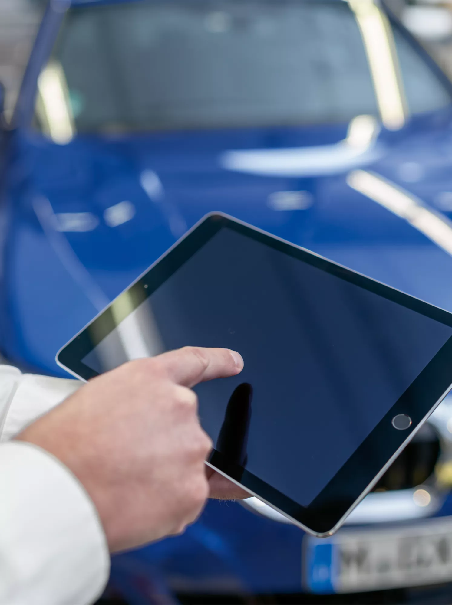 Person bedient Tablet vor blauem BMW Fahrzeug in Werkstattumgebung mit Fokus auf digitaler Fahrzeugsteuerung