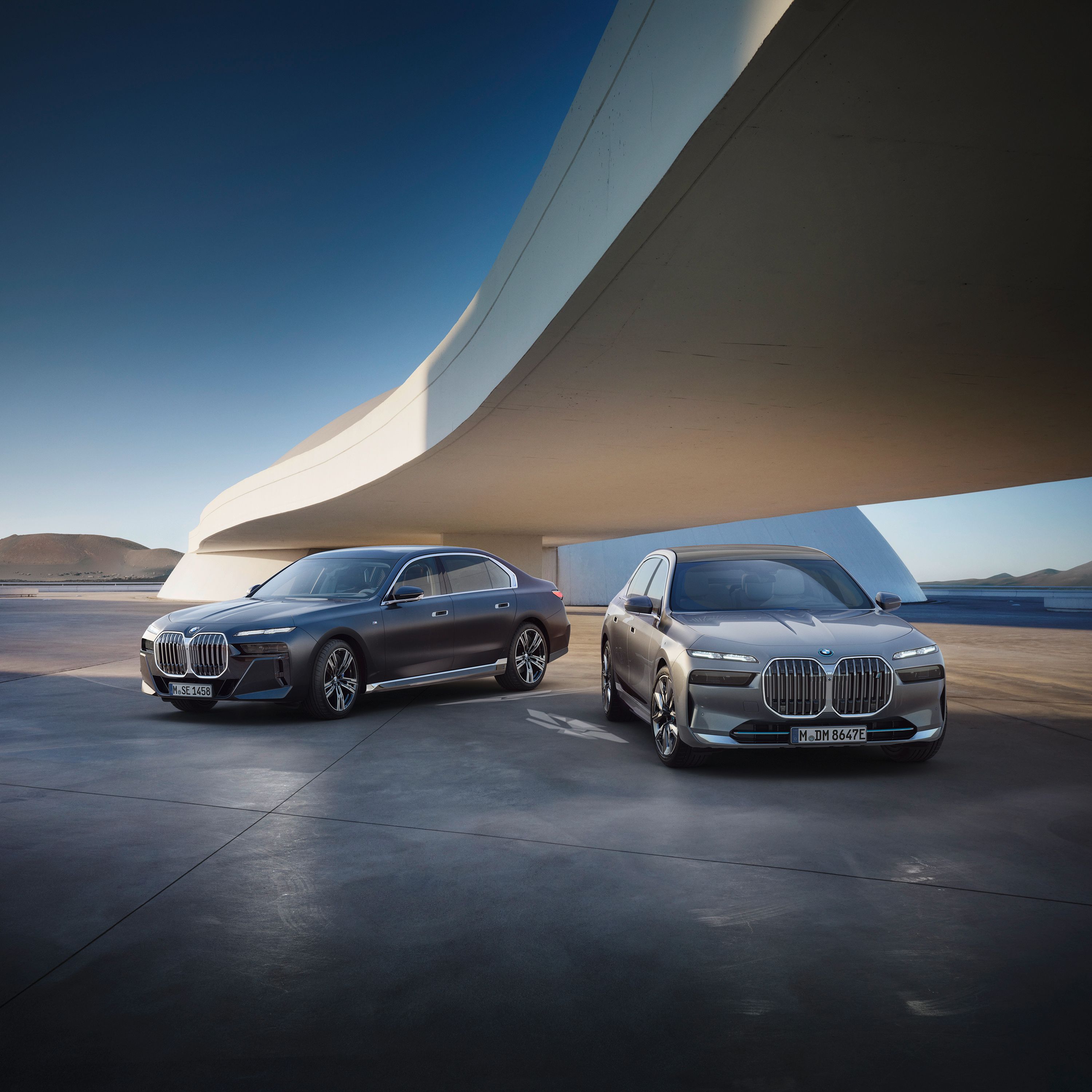 BMW i7 xDrive60 на асфальте перед башней с винтовой лестницей на фоне гор.