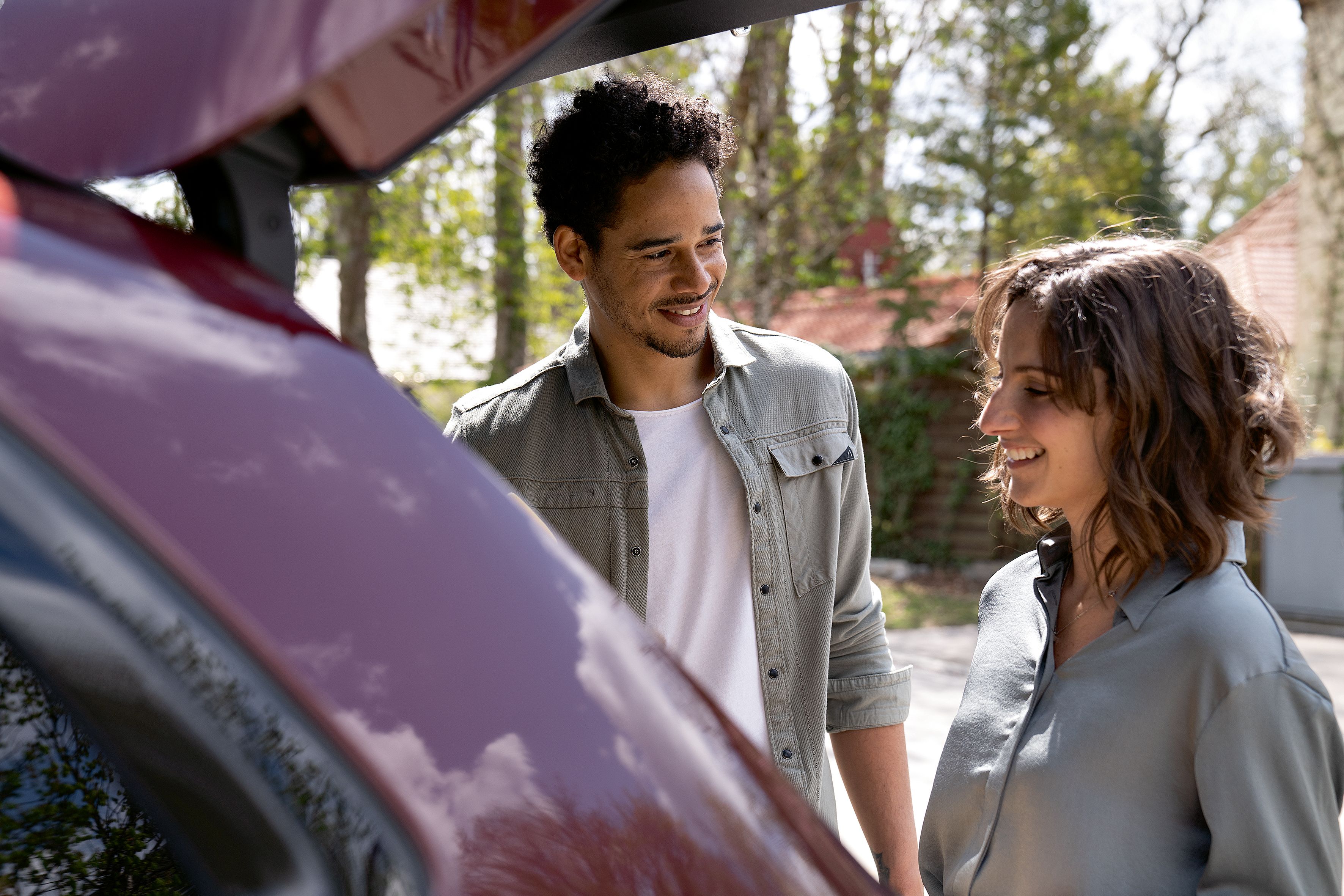 Un homme et une femme sourient devant le coffre ouvert d'une voiture rouge.