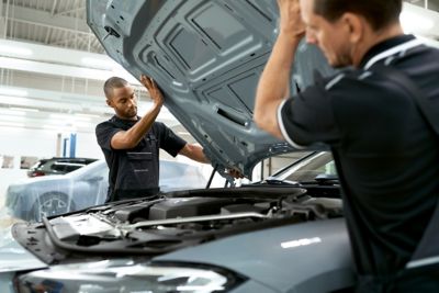 Zwei Männer in Werkstatt-Overalls heben die Motorhaube eines BMW an. Im Hintergrund ist ein abgedecktes Auto zu sehen