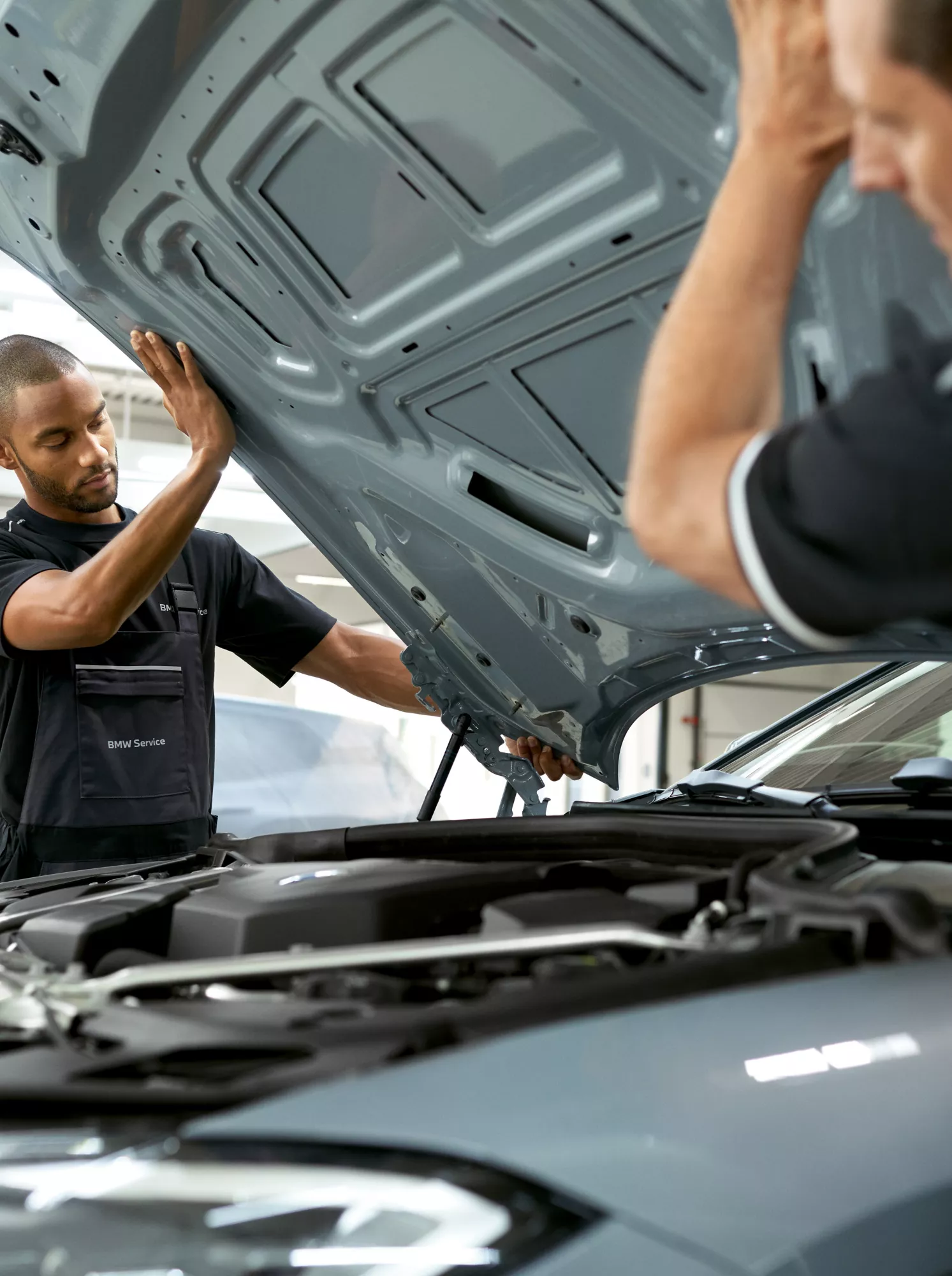 Zwei Männer in Werkstatt-Overalls heben die Motorhaube eines BMW an. Im Hintergrund ist ein abgedecktes Auto zu sehen