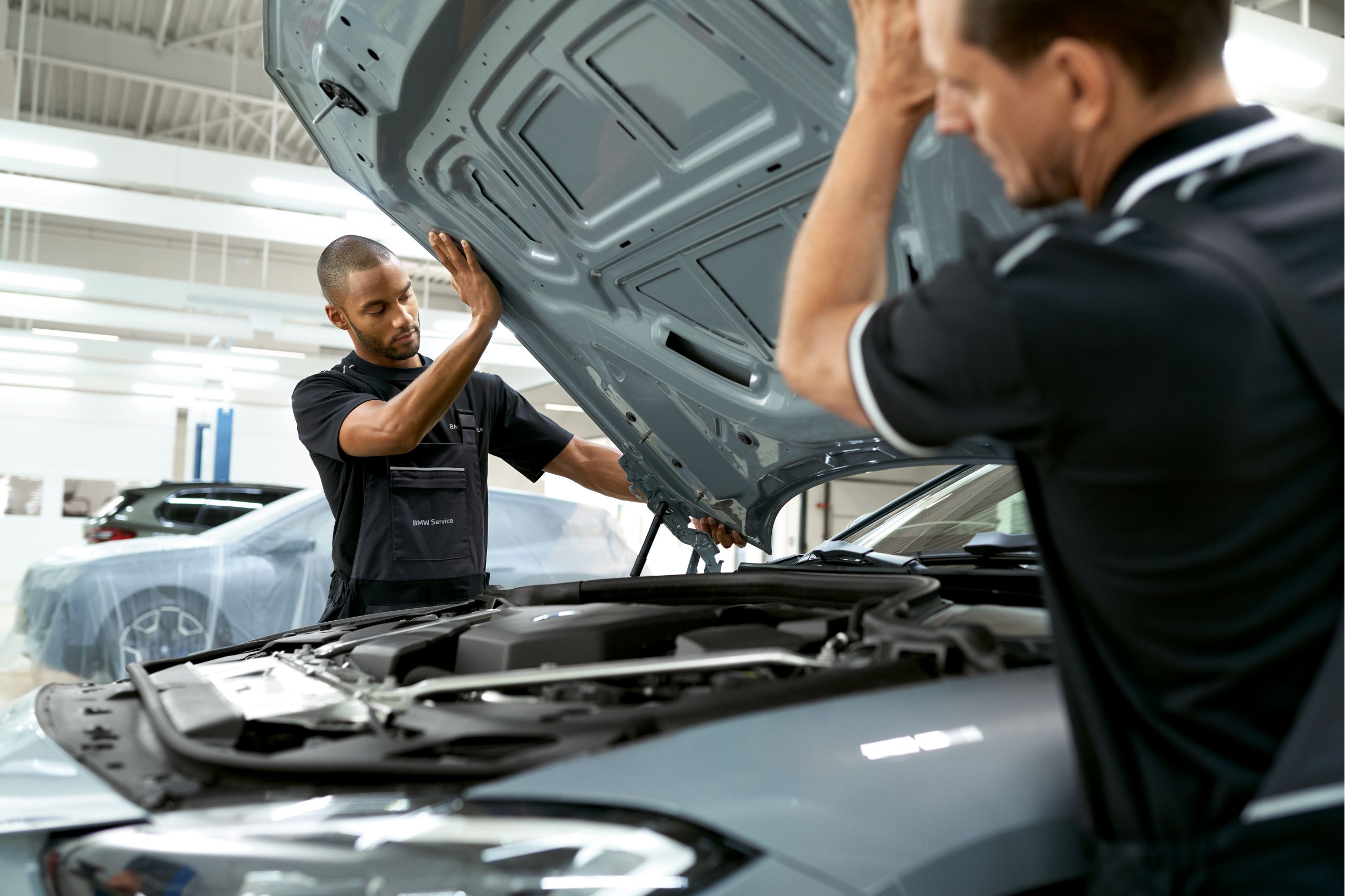 Zwei Männer in Werkstatt-Overalls heben die Motorhaube eines BMW an. Im Hintergrund ist ein abgedecktes Auto zu sehen