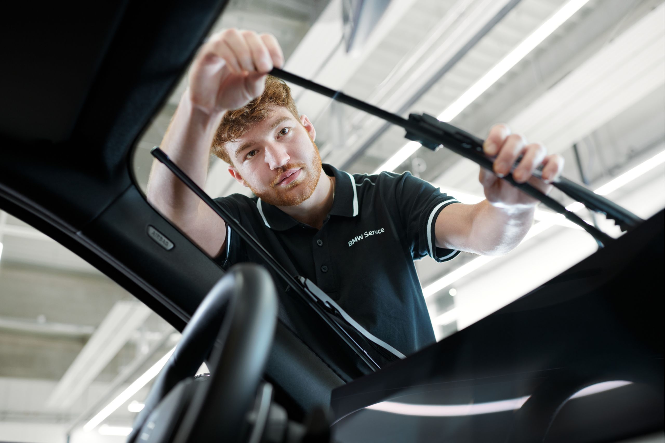 A person in a BMW Service shirt adjusts windshield wipers inside a well-lit garage. The expression is focused, conveying professionalism and care.