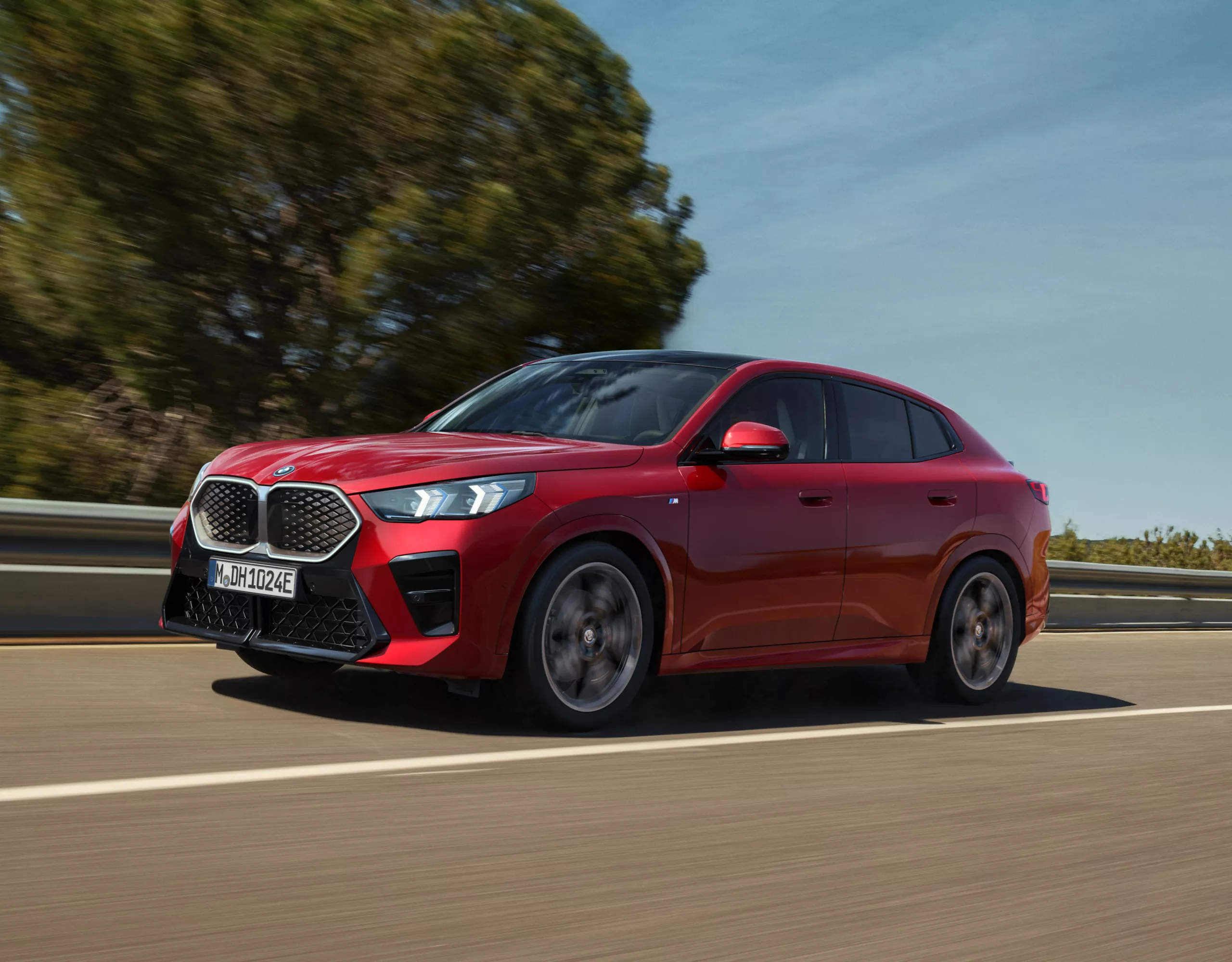 Au bord de la route, une voiture électrique BMW se recharge sur une borne publique. Une personne appuyée contre la BMW regarde son smartphone