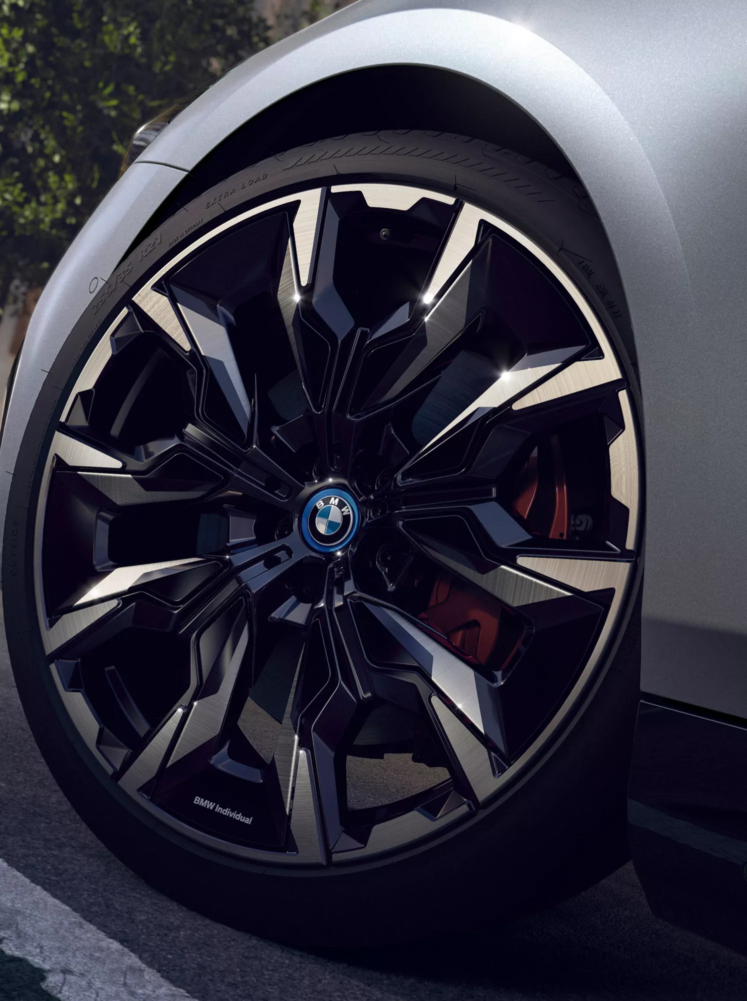 Im Fokus ist eine BMW Felge die an einem silbernen Auto befestigt ist, dass über eine Straße fährt