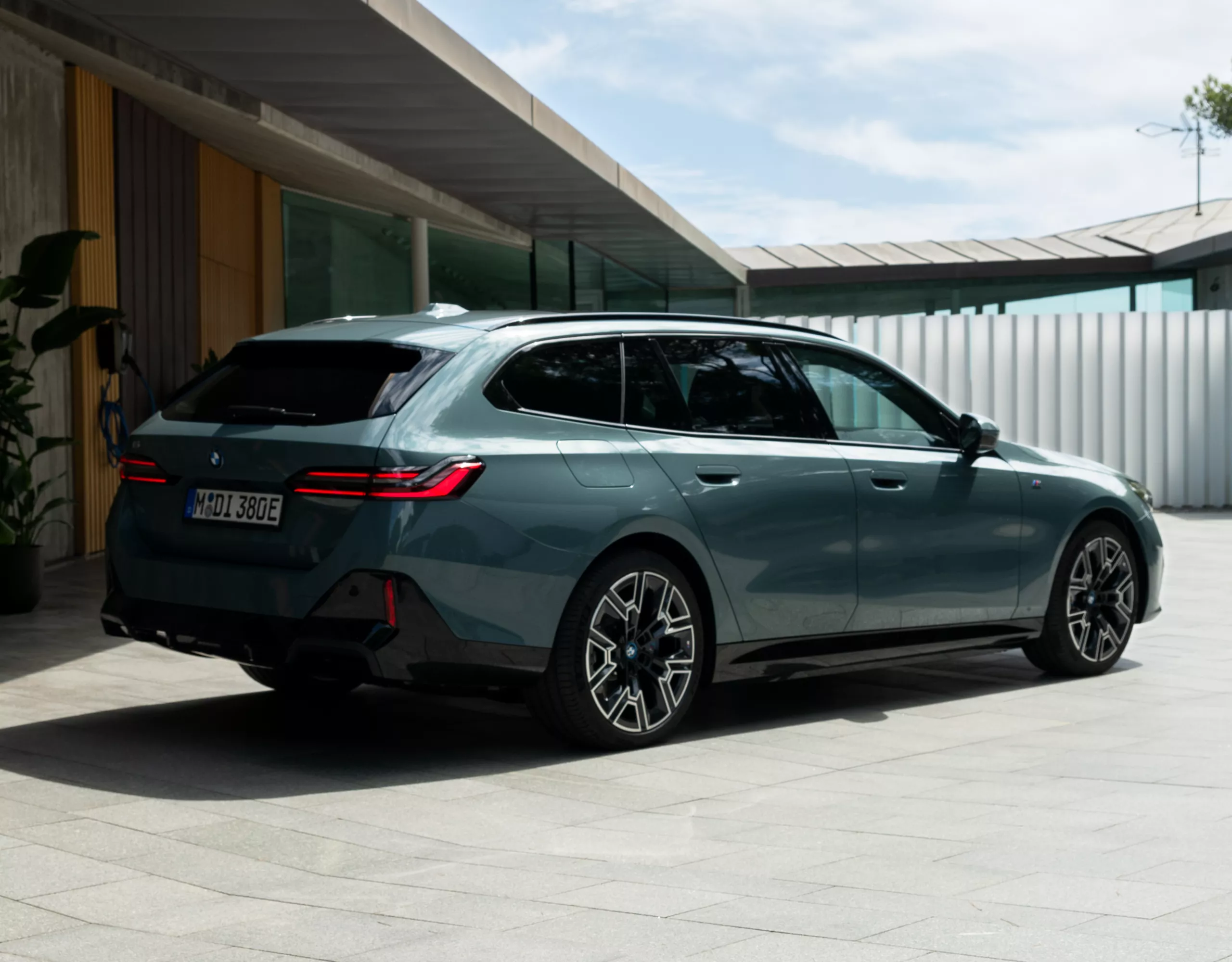 Une BMW Touring grise est garée devant une maison moderne faite de béton et d'éléments en bois.