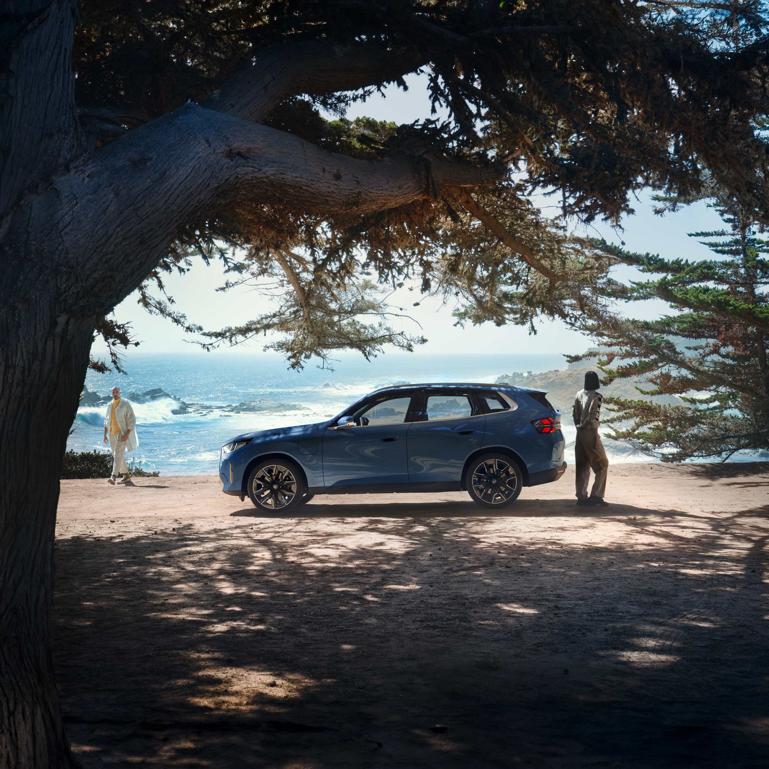 BMW iX2 all-electric vehicle in Brooklyn Grey color driving on a road at sunset with an ocean view in the background.