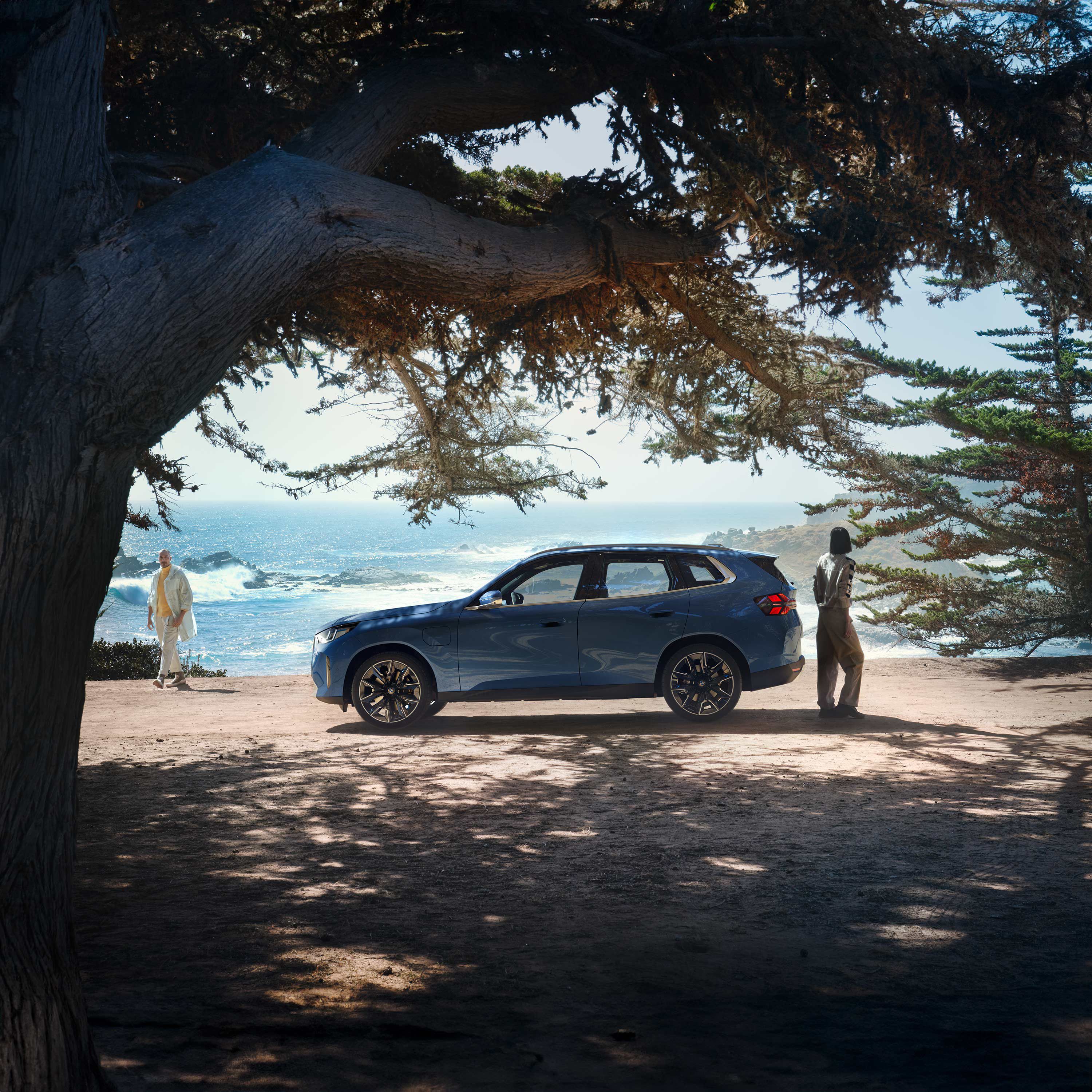 BMW iX2 all-electric vehicle in Brooklyn Grey color driving on a road at sunset with an ocean view in the background.