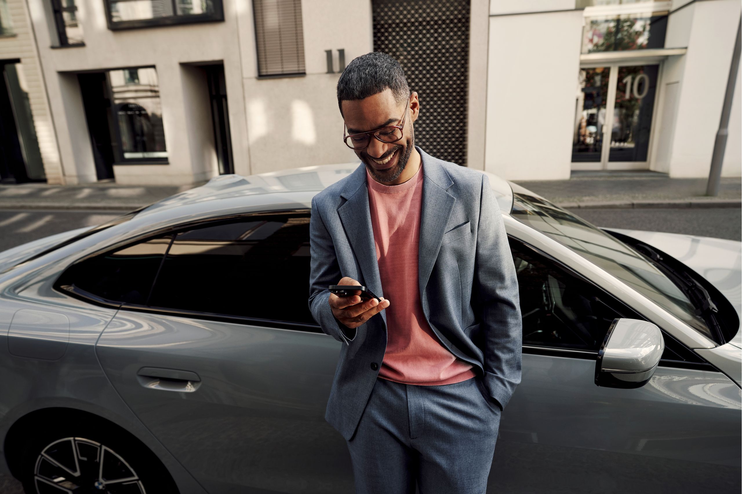 A driver leans against the passenger door of his BMW while looking down at his smartphone