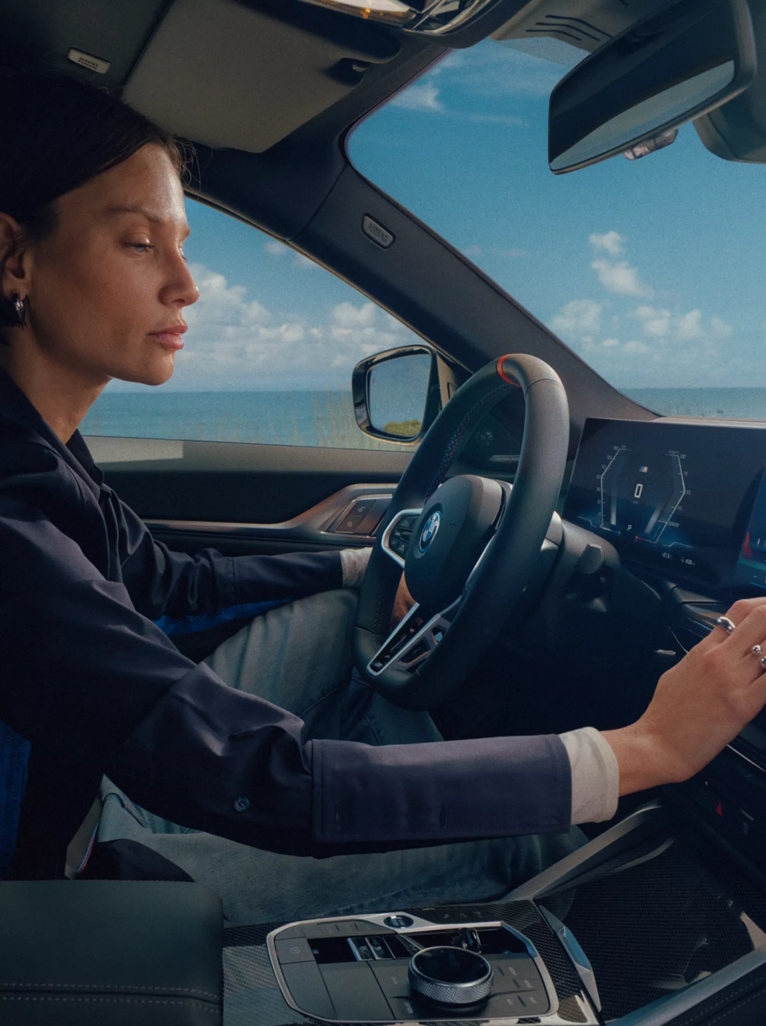 BMW Fahrerin bedient das hochmoderne BMW Cockpit mit Curved Display vor einer malerischen Küstenlandschaft.