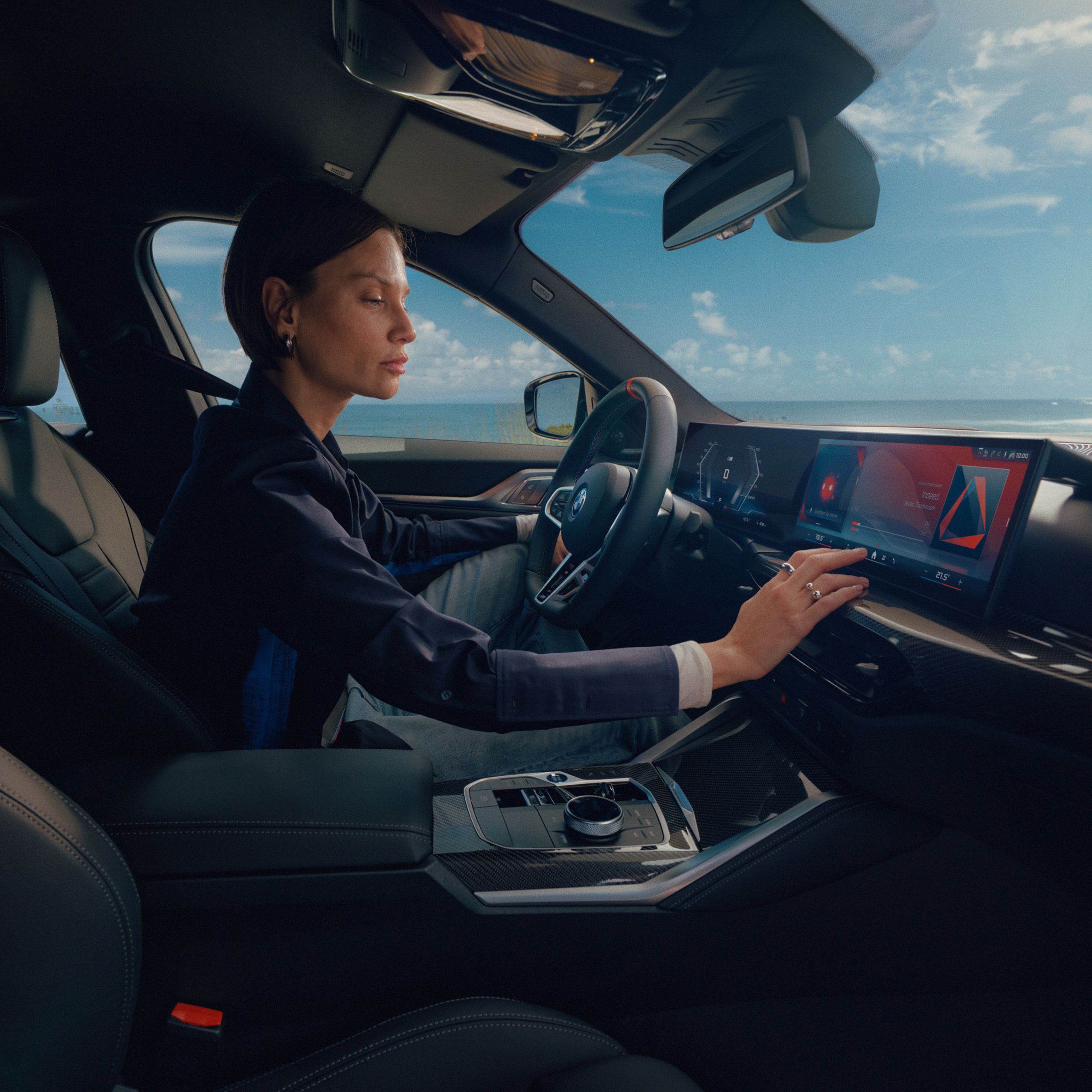 BMW Fahrerin bedient das hochmoderne BMW Cockpit mit Curved Display vor einer malerischen Küstenlandschaft.