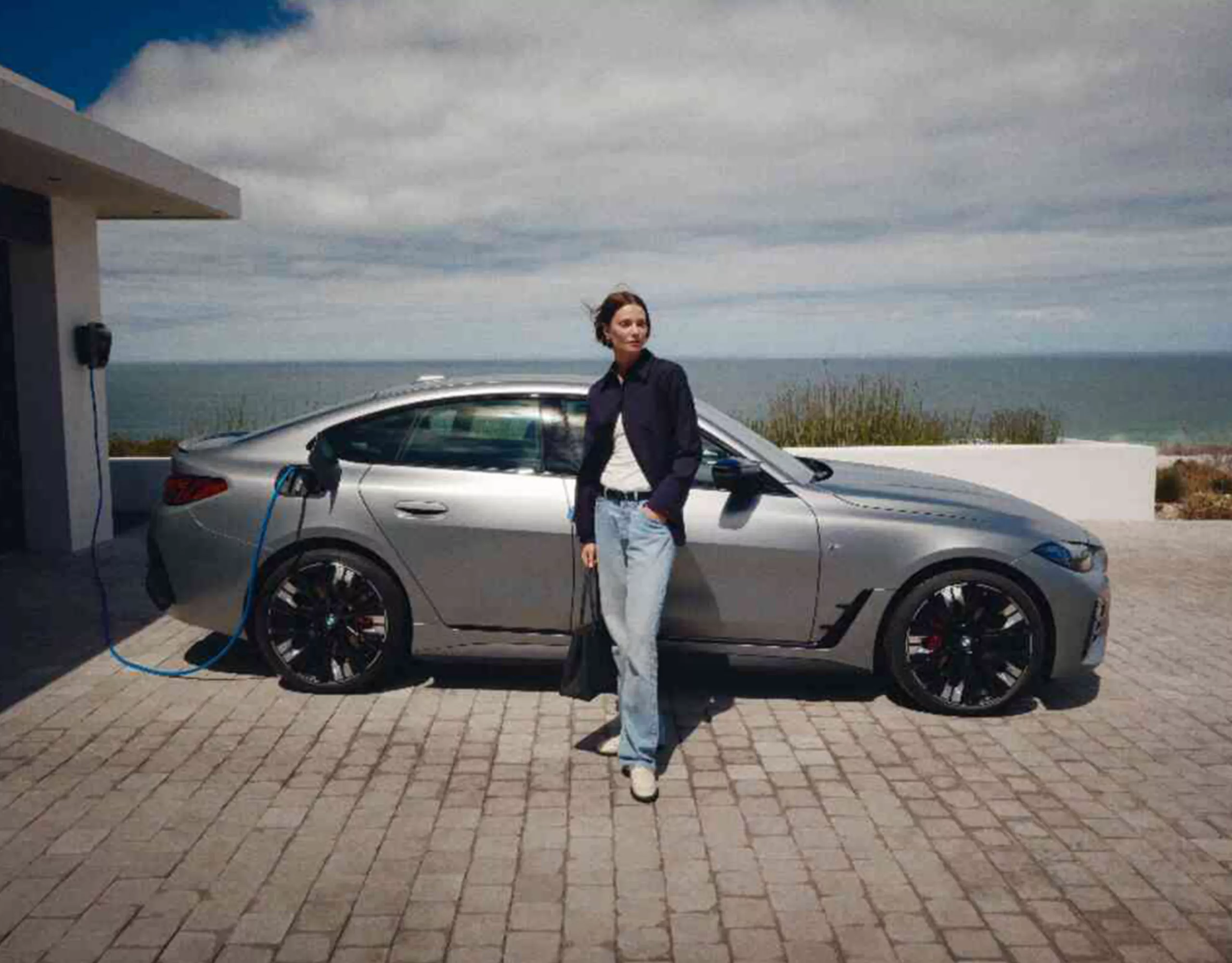 A grey BMW and a man are standing in front of a modern glass building, while the BMW is being charged at a public charging spot