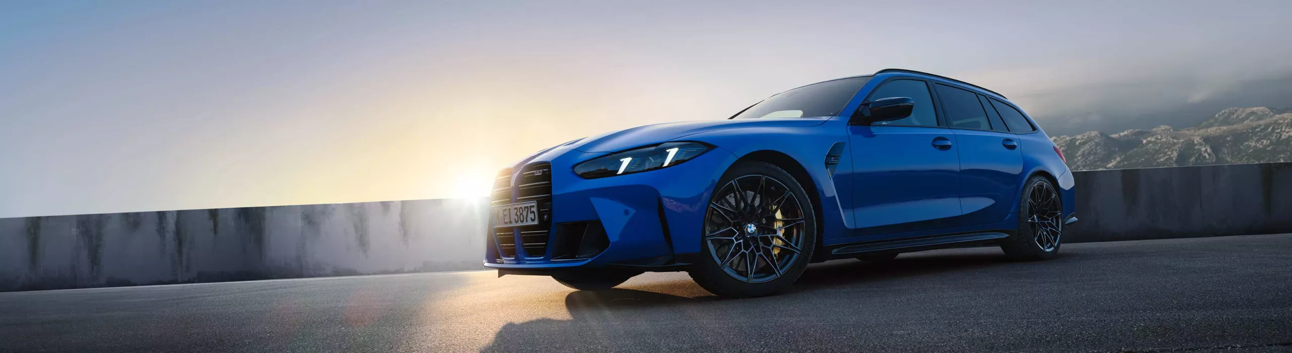 BMW M3 Touring in blue parked at sunrise on a scenic mountain road with a dramatic sky
