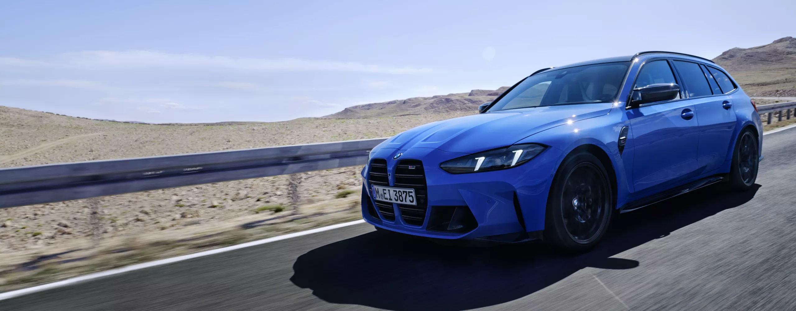 Blauer BMW M3 Touring fährt auf einer leeren Straße durch eine wüstenähnliche Landschaft unter blauem Himmel
