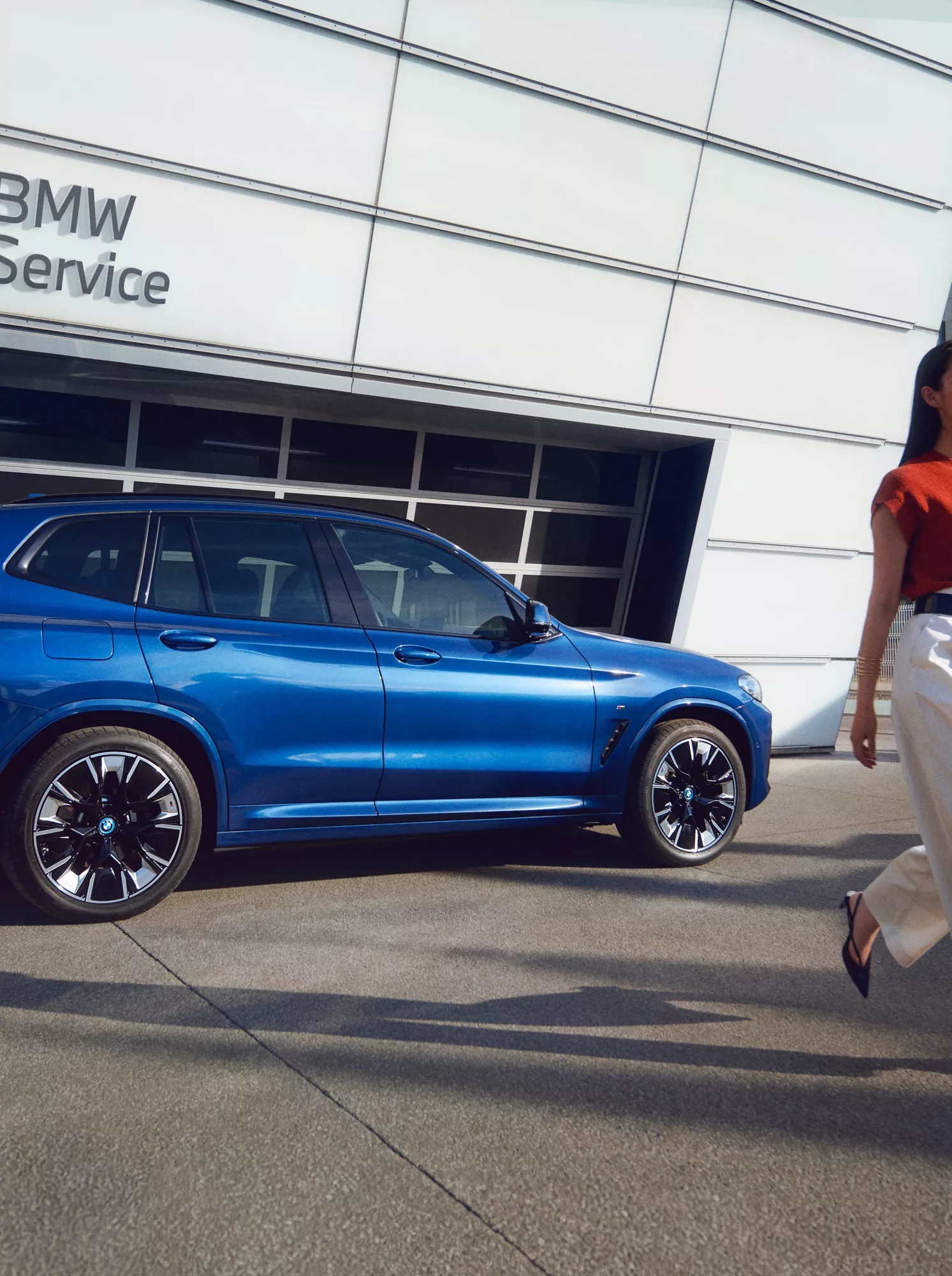 Ein blauer SUV von BMW steht vor den Garagentoren eines BMW Service Betriebs. Eine Frau geht von dem Fahrzeug weg.
