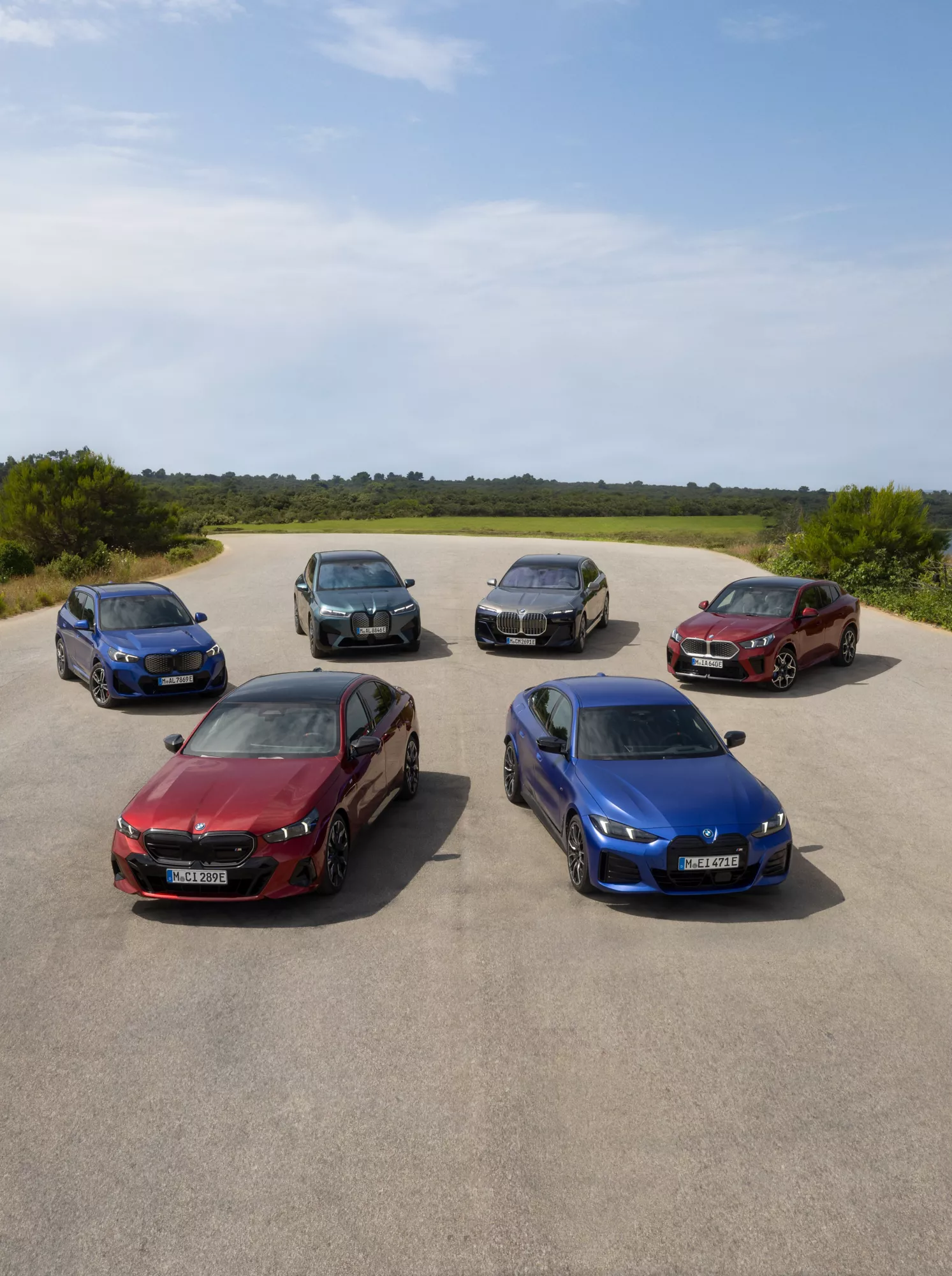 Sechs BMW Modelle stehen auf einer Straße in grüner Landschaft. Im Vordergrund ist ein rotes und ein blaues Modell zu sehen