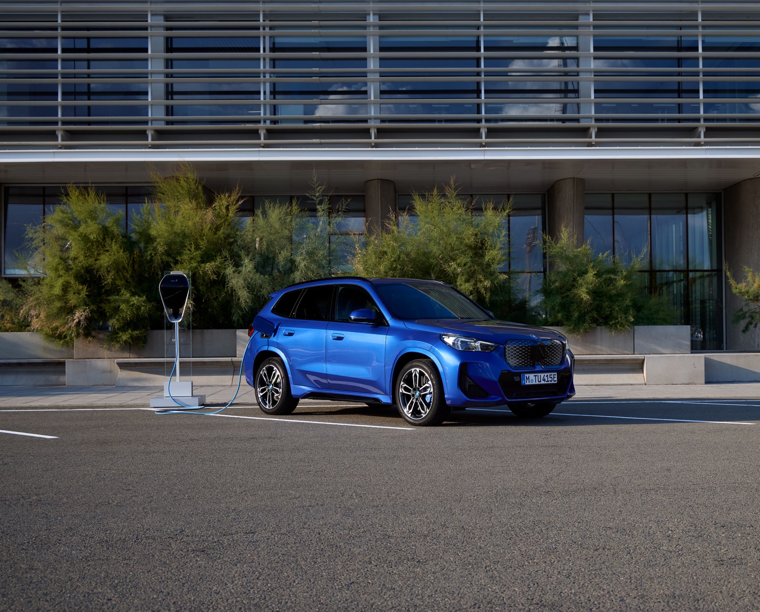 Obrázek znázorňující vůz BMW iX1 v modré barvě stojící na parkovišti připojený k nabíječce.