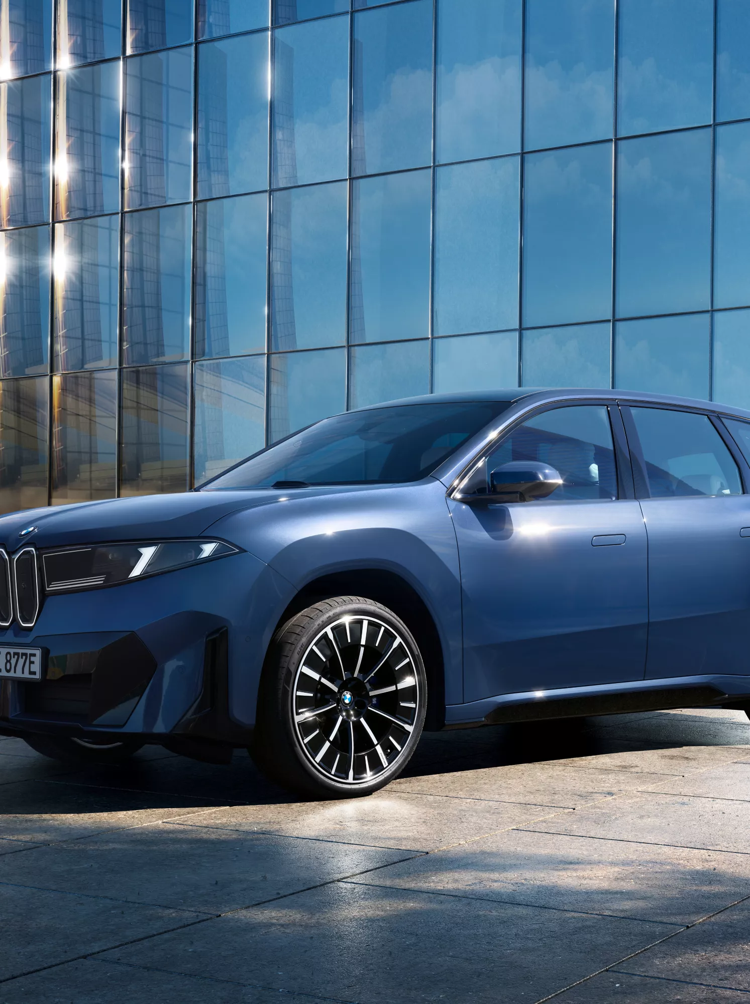 A sleek blue SUV is parked on a tiled surface beside a reflective glass building. Sunlight glistens on the facade, creating a modern and sophisticated ambiance.