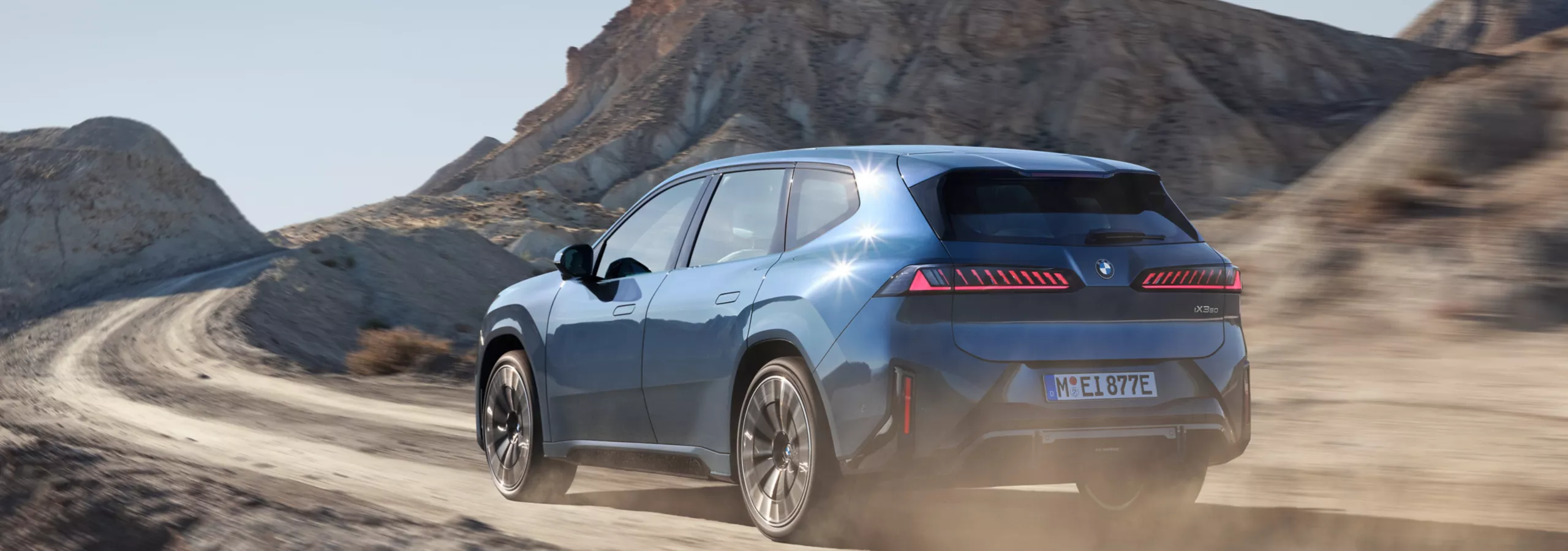 BMW iX3 fährt auf kurviger Schotterstraße durch felsige Landschaft, Heckansicht mit Staubaufwirbelung und LED-Rückleuchten
