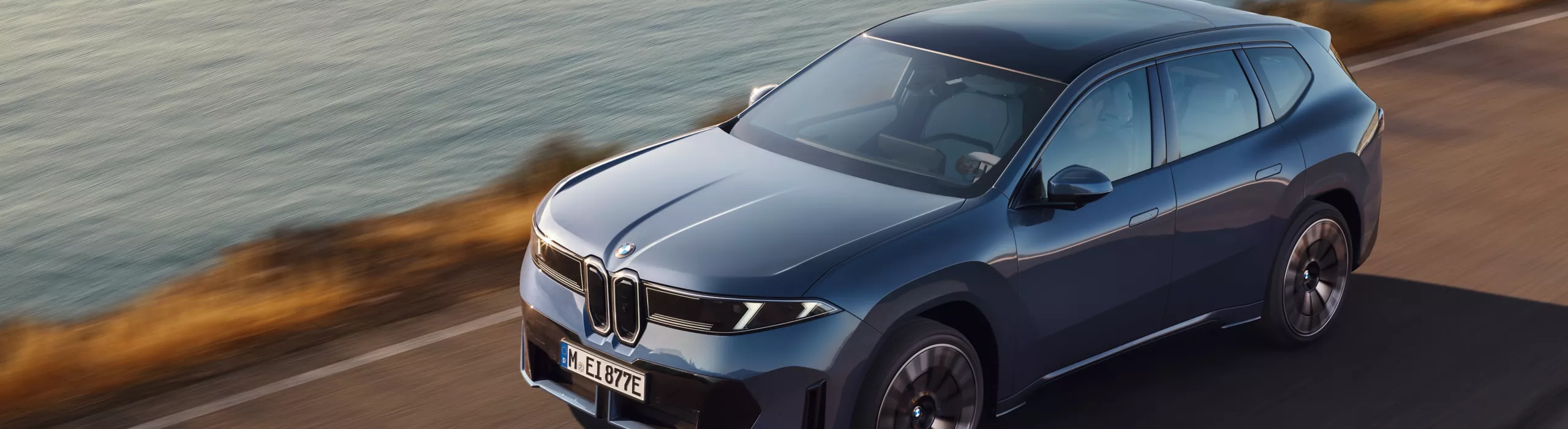 BMW iX3 fährt entlang einer Küstenstraße bei Sonnenuntergang, dynamische Seitenansicht mit Meer und Felsen