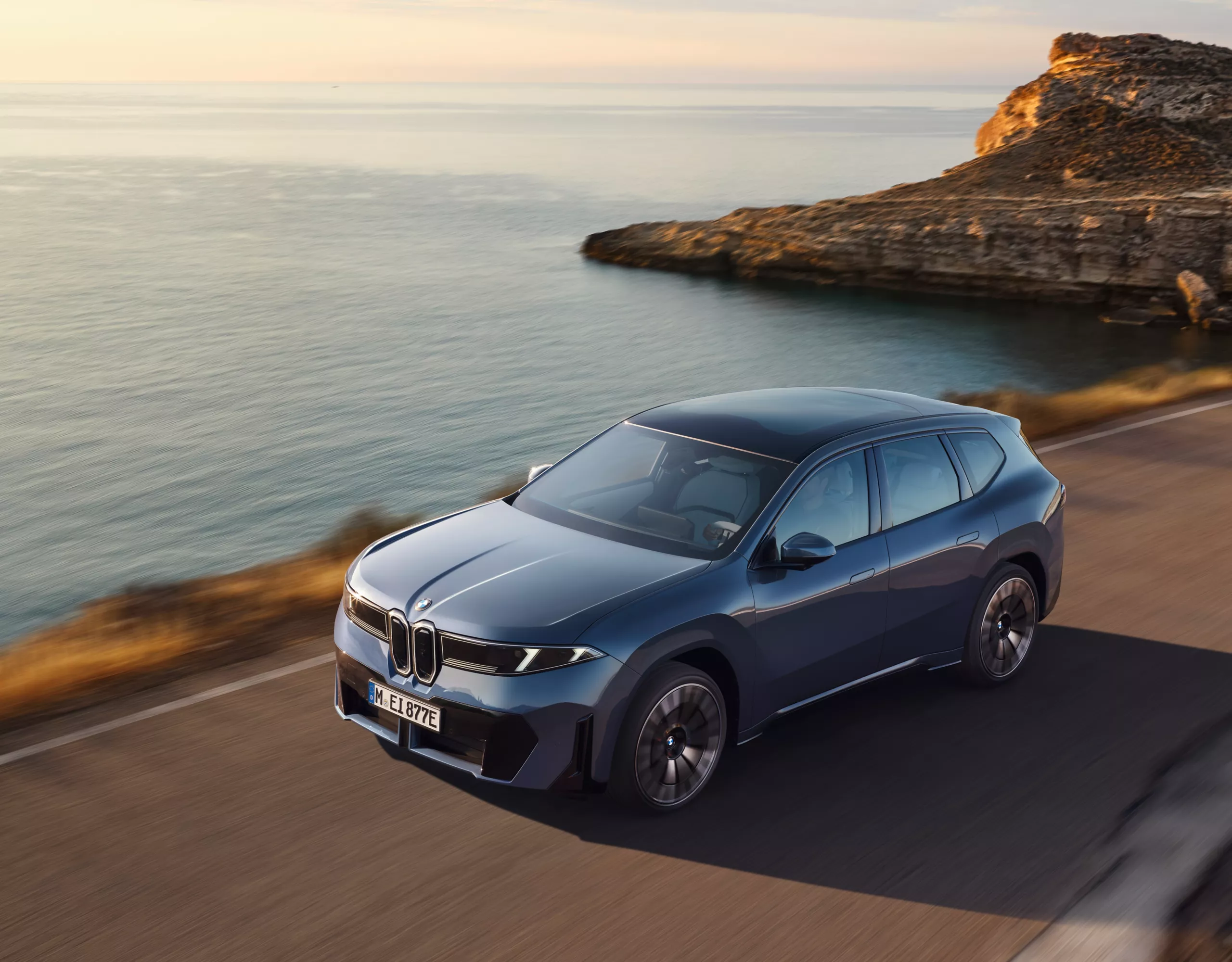 BMW iX3 fährt entlang einer Küstenstraße bei Sonnenuntergang, dynamische Seitenansicht mit Meer und Felsen