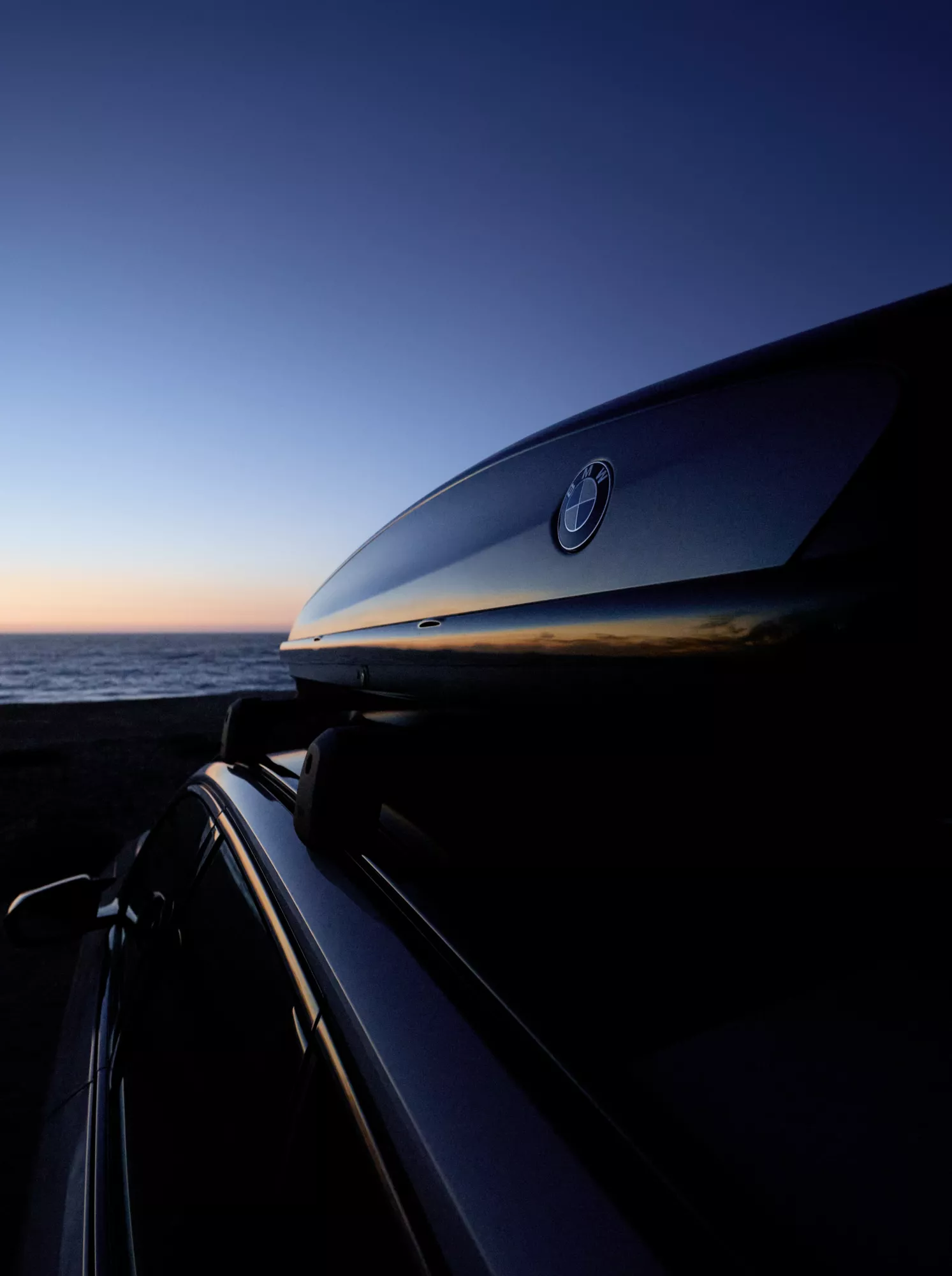 A BMW with a roof box stands in the sunshine in an open field, a man leans against the vehicle, a woman looks into the distance