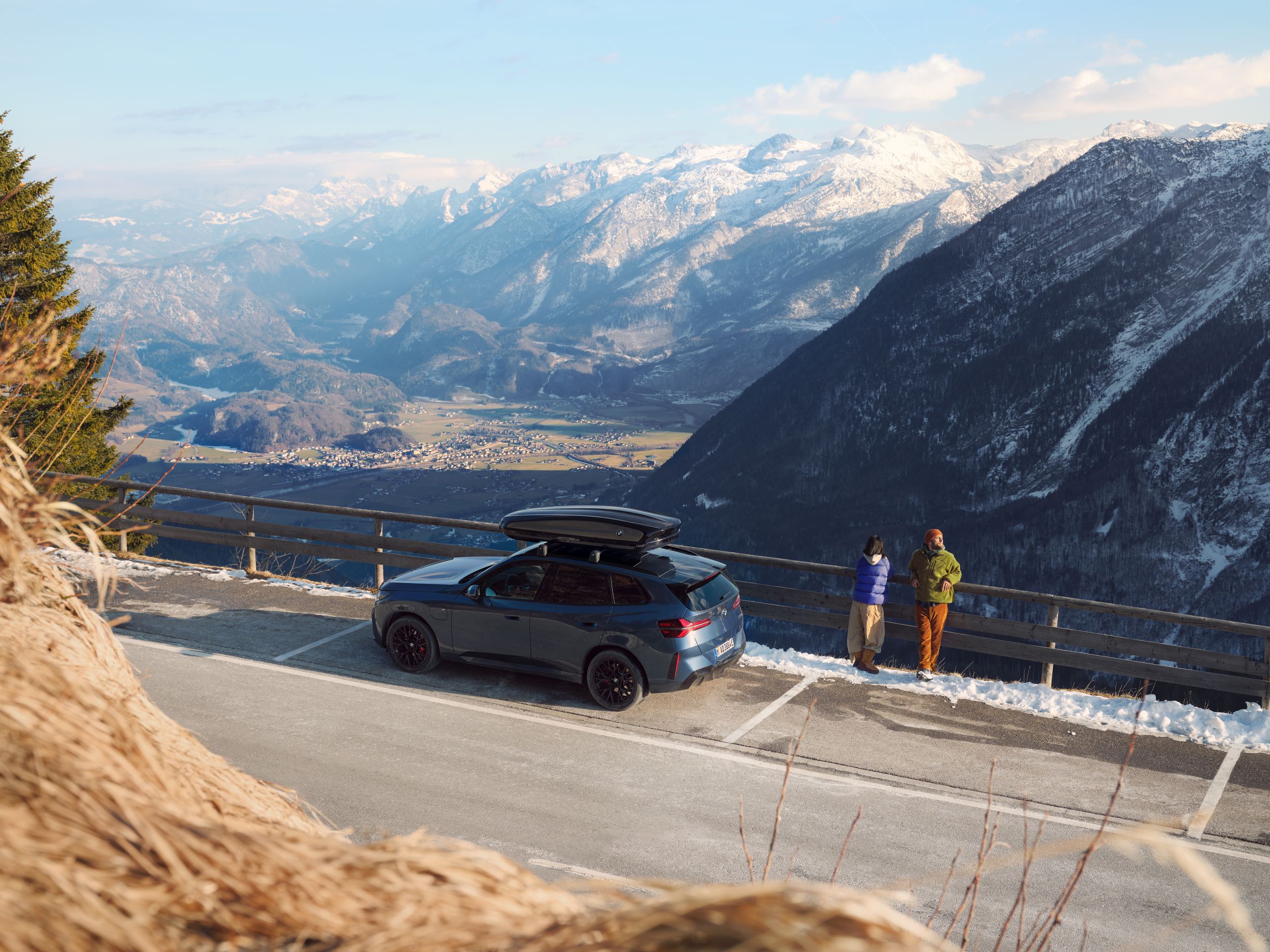 Une BMW avec un coffre de toit est stationnée sur une route de montagne tandis que deux personnes regardent les vallées et les montagnes