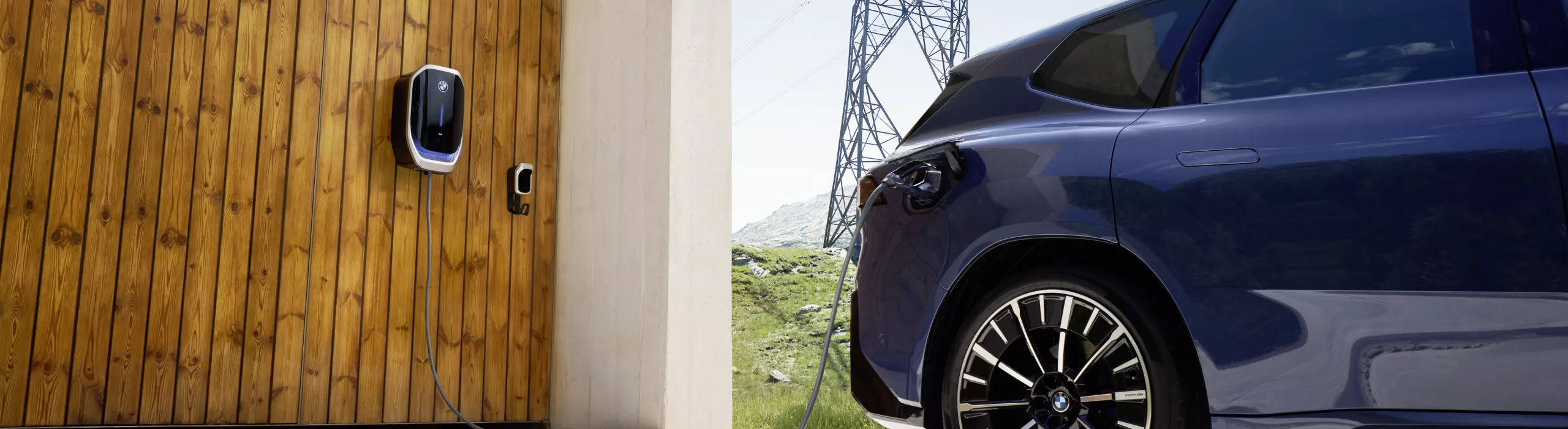 Elektroauto lädt an einer Wallbox an einer Holzfassade, im Hintergrund Strommasten und hügelige Landschaft