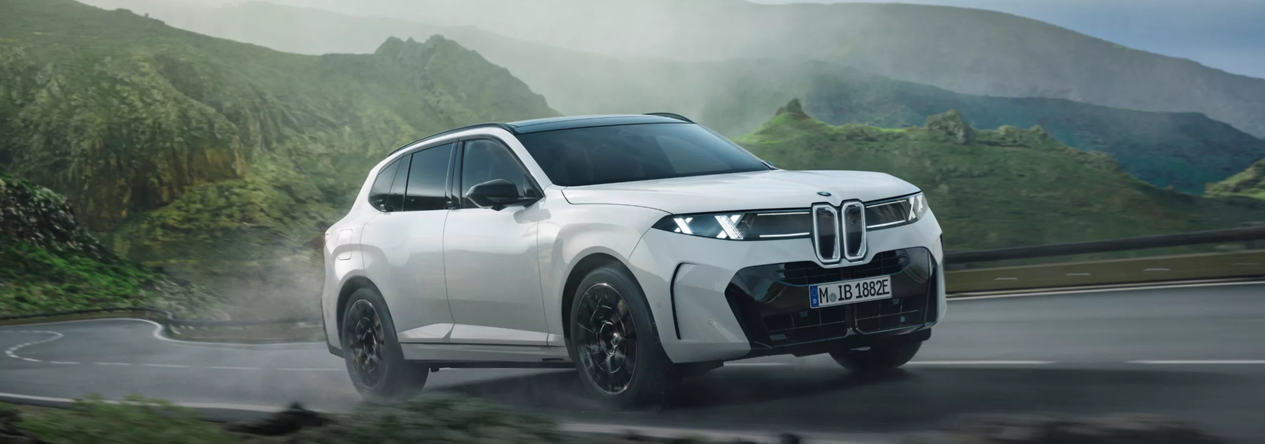 BMW X5 branco em movimento numa estrada de montanha, com paisagem verde, céu nublado e mar ao fundo