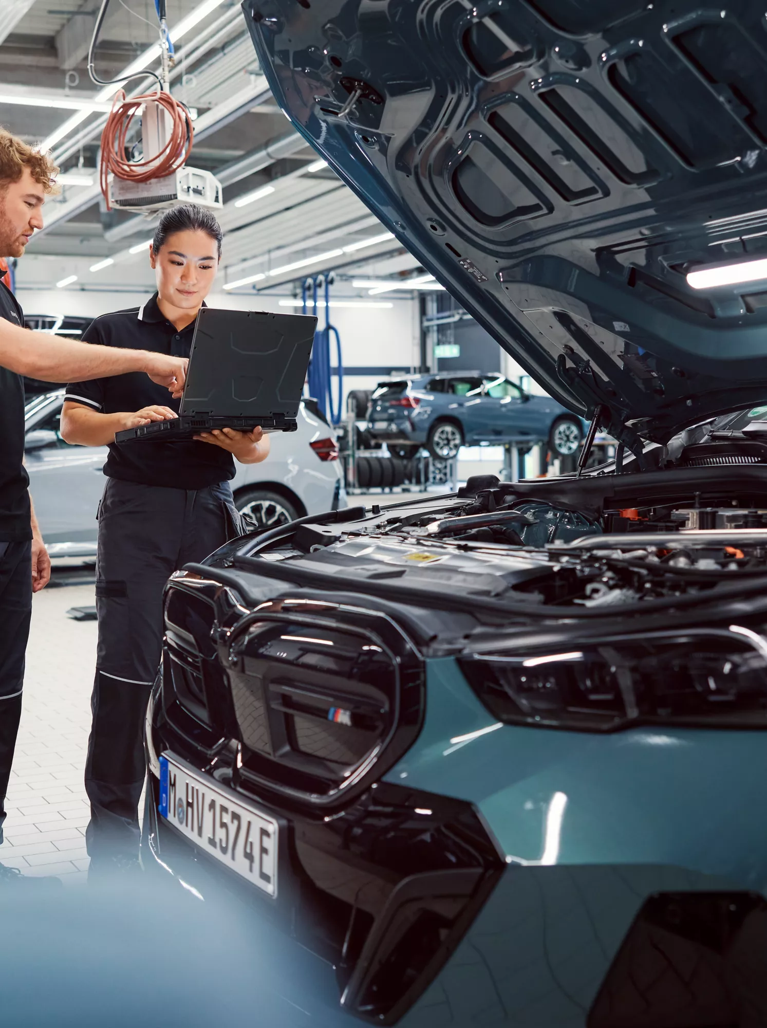 Dois técnicos BMW Service a consultar um computador portátil junto a um BMW com o capô aberto, dentro de uma oficina