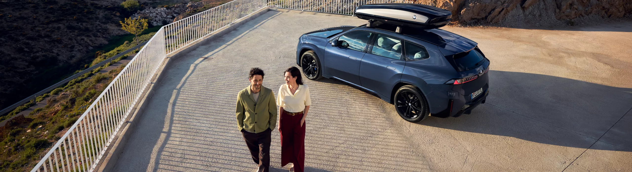 Une BMW avec un coffre de toit est stationnée sur une route de montagne tandis que deux personnes regardent les vallées et les montagnes