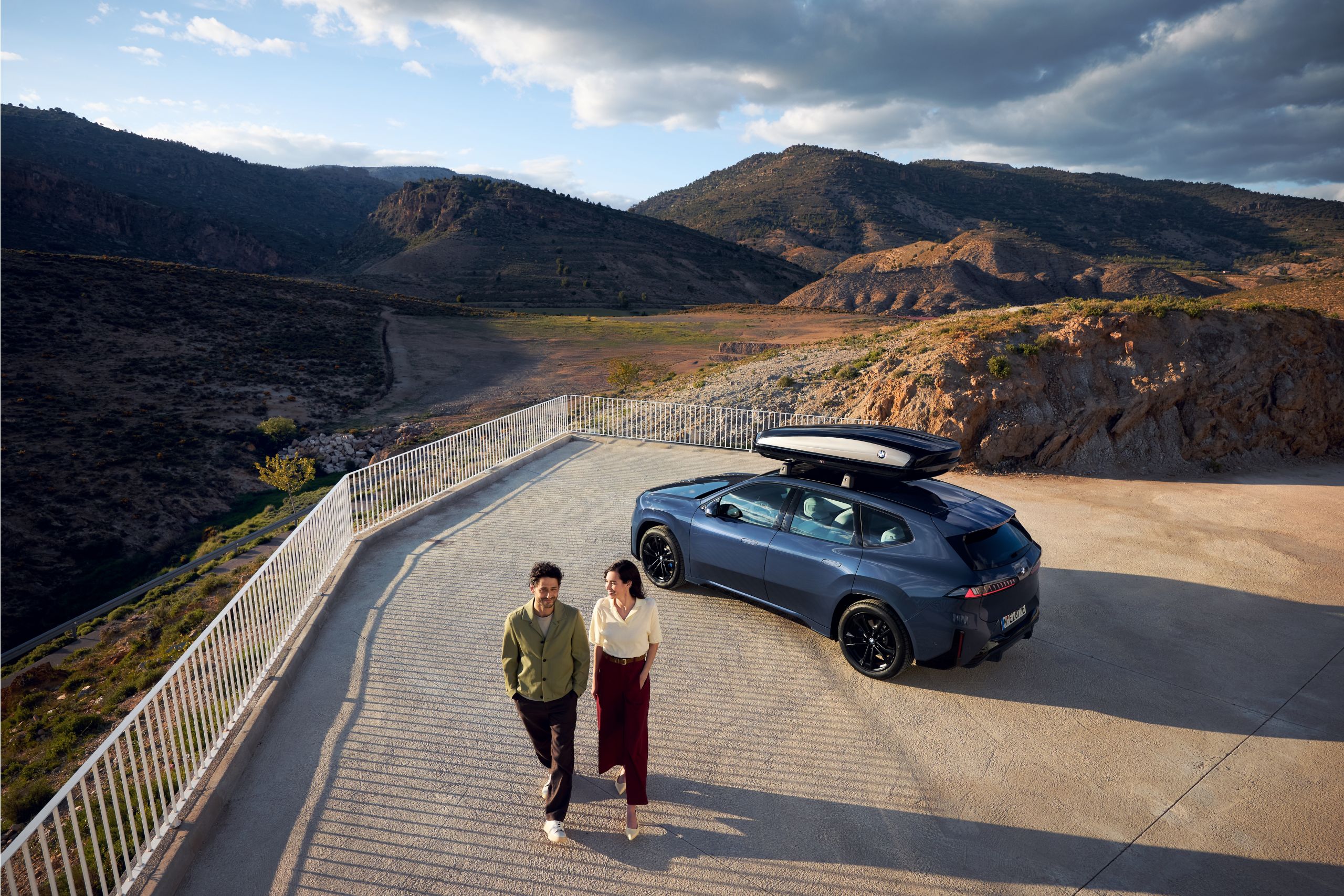 Une BMW avec un coffre de toit est stationnée sur une route de montagne tandis que deux personnes regardent les vallées et les montagnes