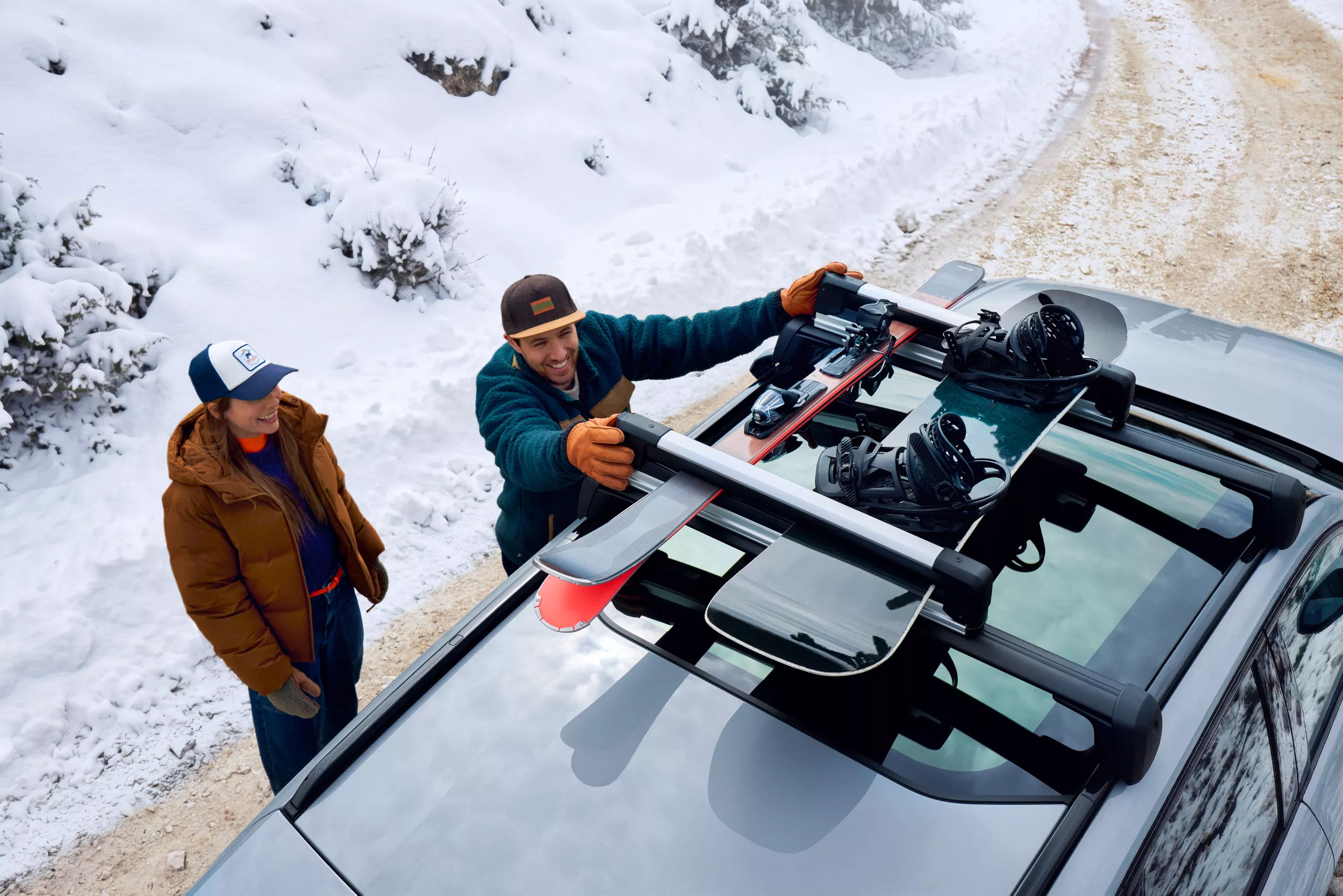 Homem a colocar equipamento de esqui no tejadilho de um BMW, com uma mulher ao lado, numa estrada coberta de neve