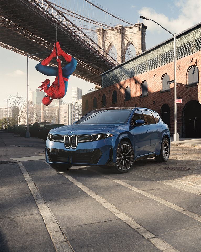 Spiderman dangles upside down in front of the iX3, which is parked in an abandoned city-scape in front of a historic theater.