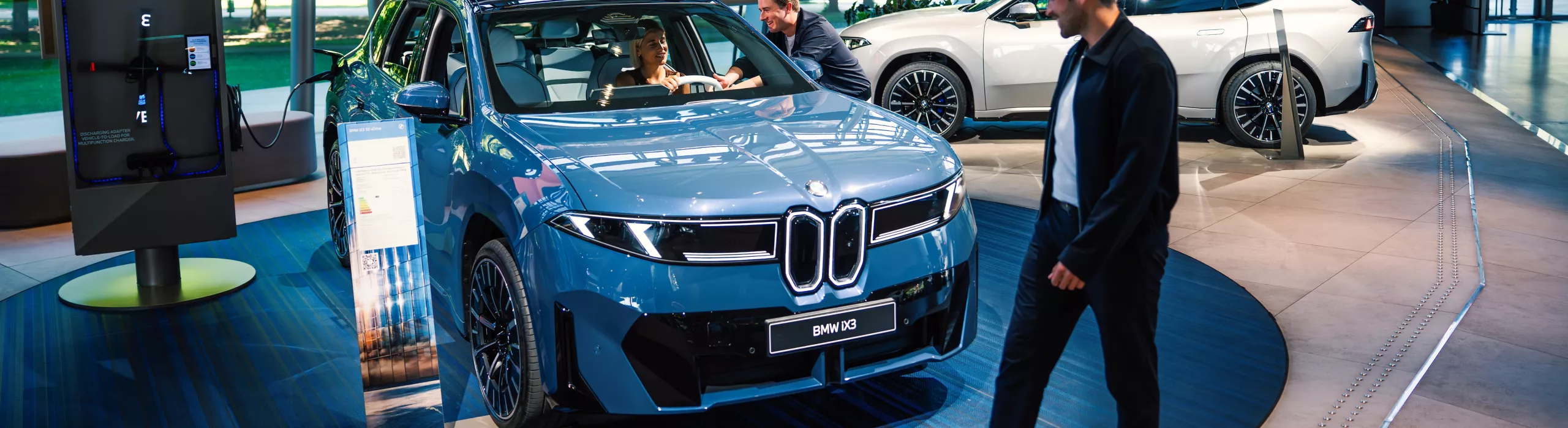 BMW iX3 in der BMW Welt in München; eine Person sitzt im Fahrzeug, während ein Mitarbeitender Funktionen erklärt, im Hintergrund ein großer digitaler Bildschirm und ein weiteres BMW Modell.