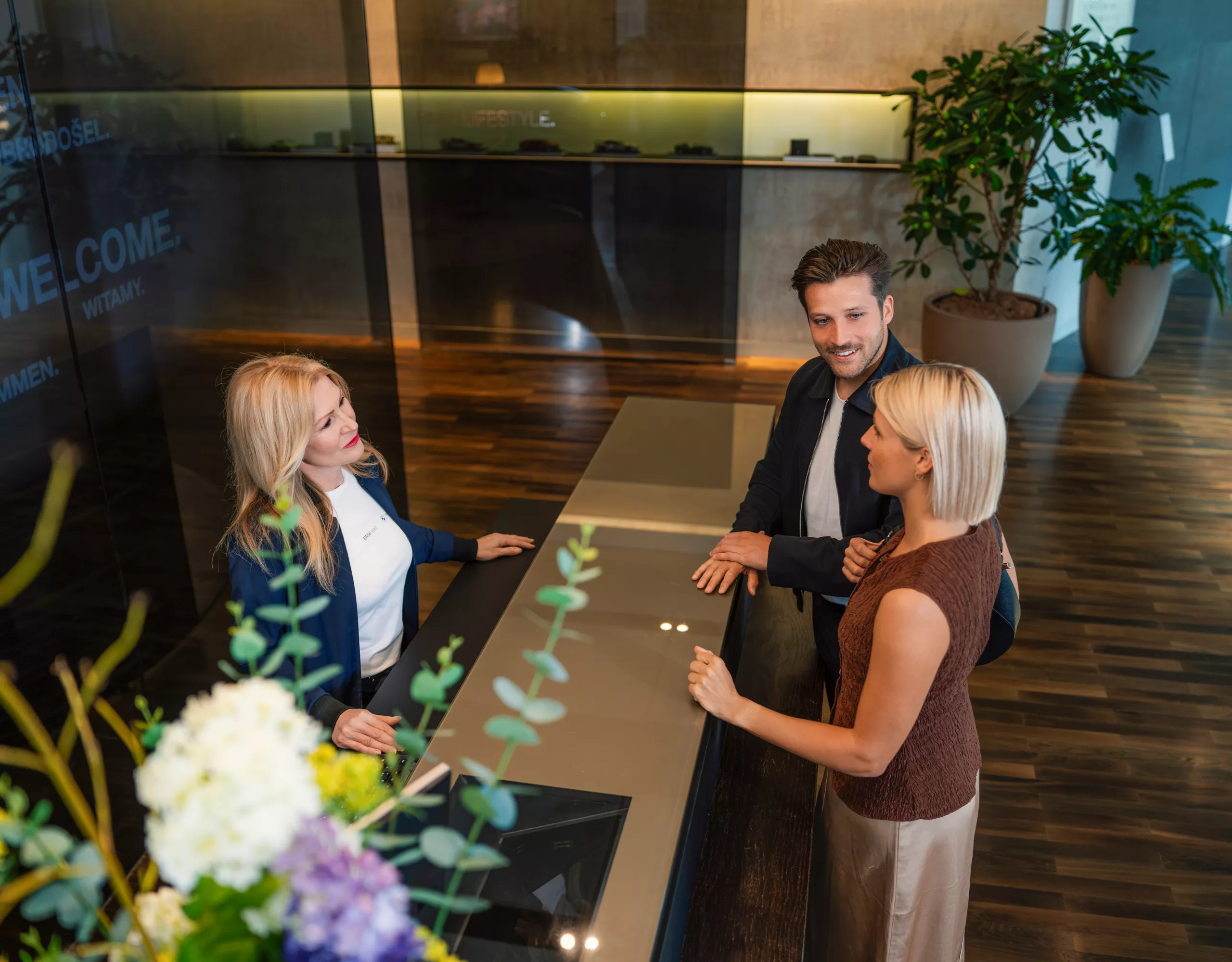 A staff member welcomes two visitors at the counter at the Check-In area.