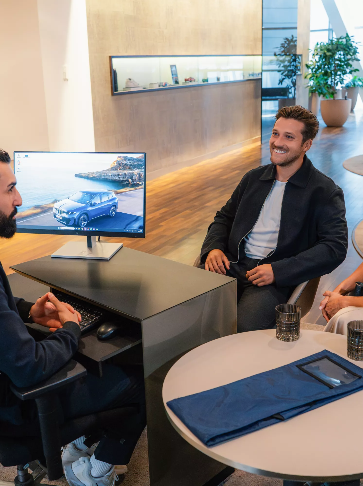 BMW Genius consults two visitors at BMW Welt, presenting vehicle information on a screen.