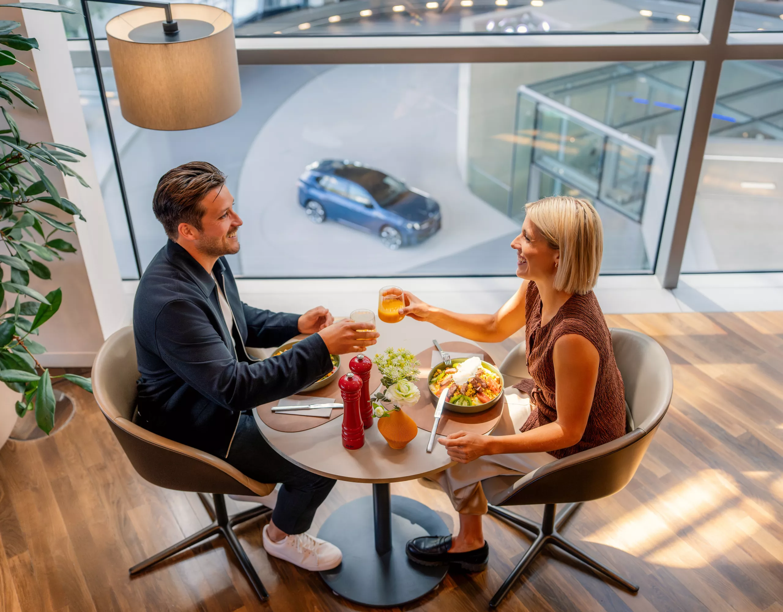 A couple sits in the BMW Welt Premium Lounge, toasting with drinks while enjoying a meal together.