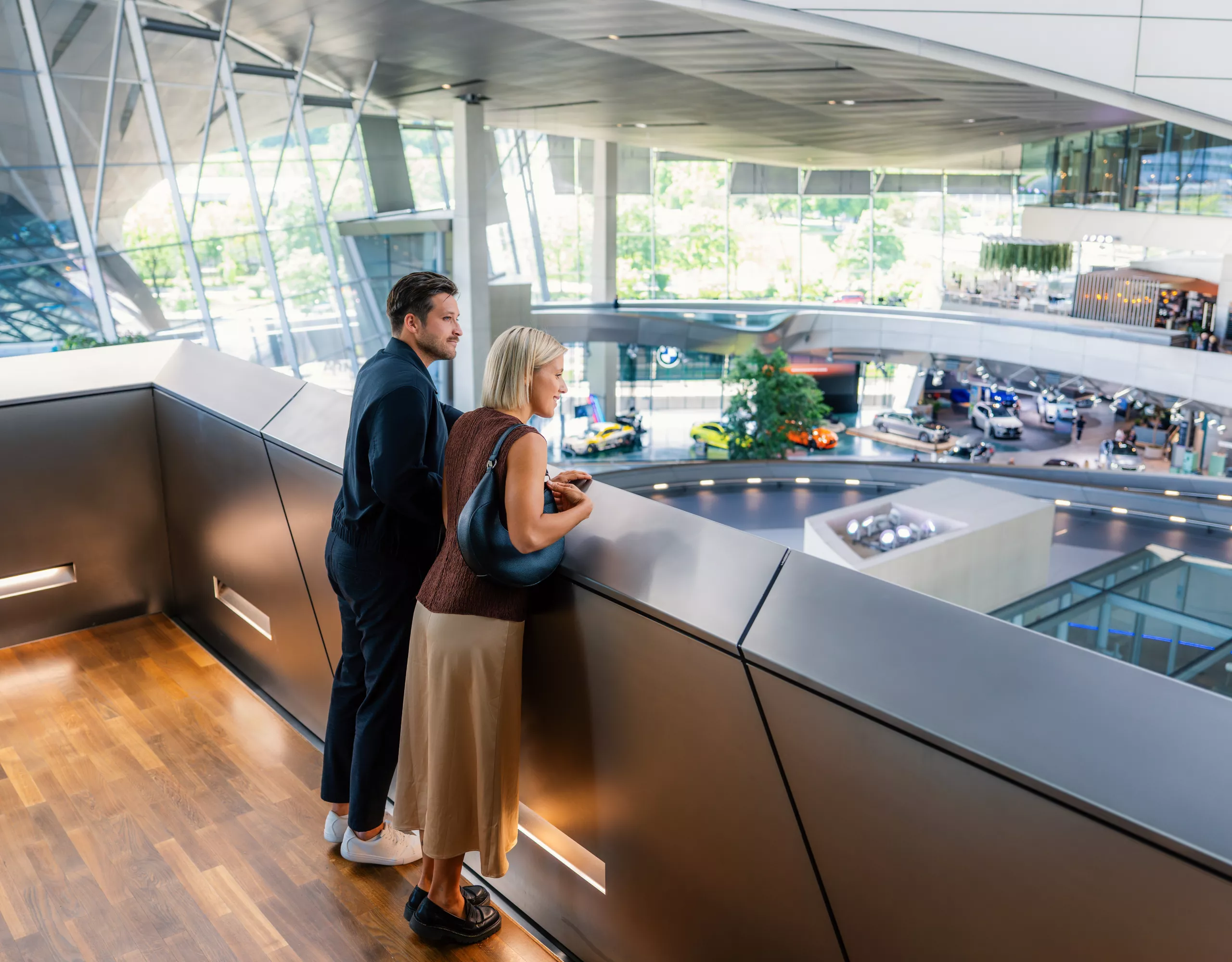 The couple stands on an upper level at BMW Welt, overlooking the open vehicle exhibition with several displayed models.