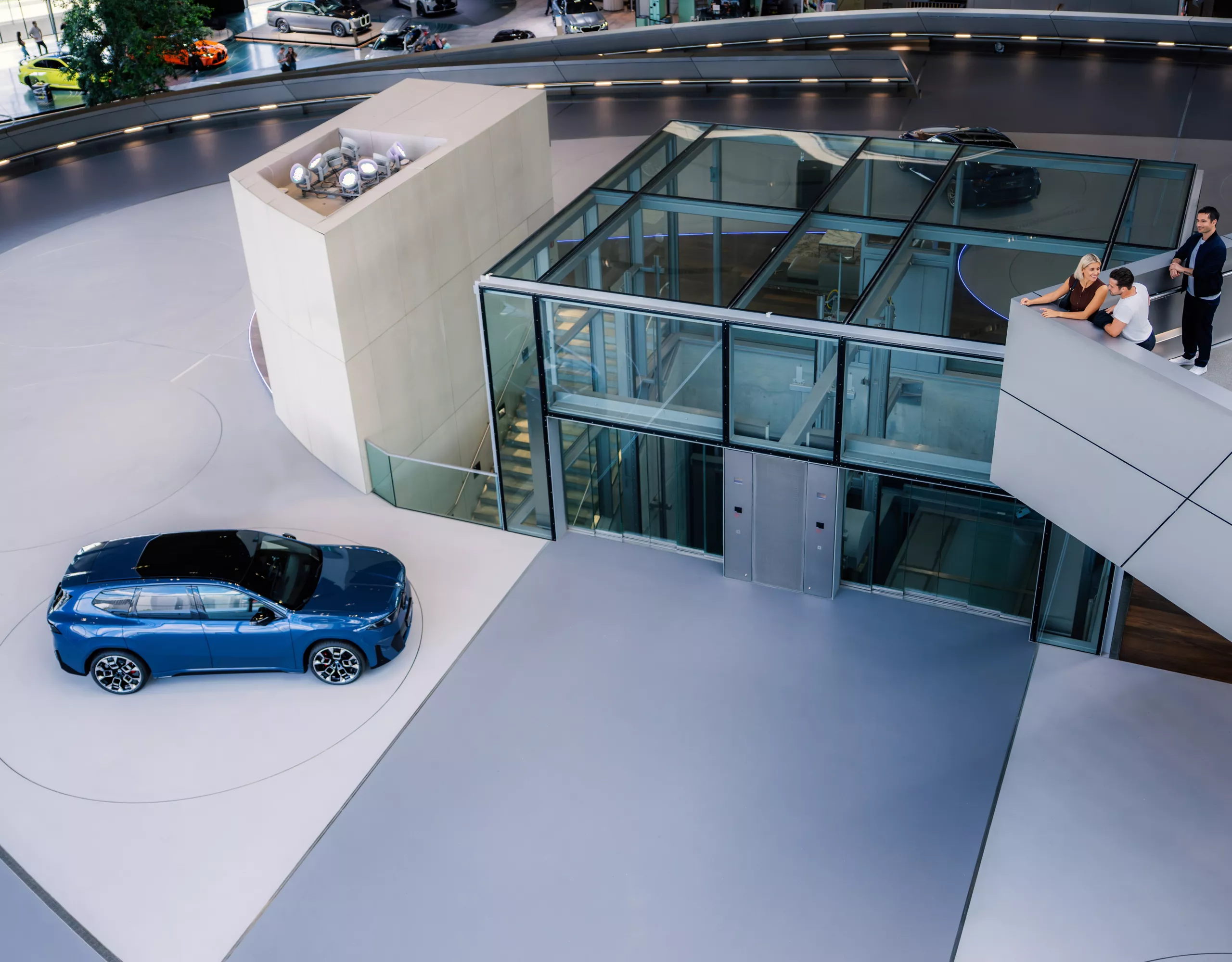 BMW iX3 in blue showcased in BMW Welt’s futuristic exhibition space, seen from above with glass elevator structure and visitors observing from a balcony.