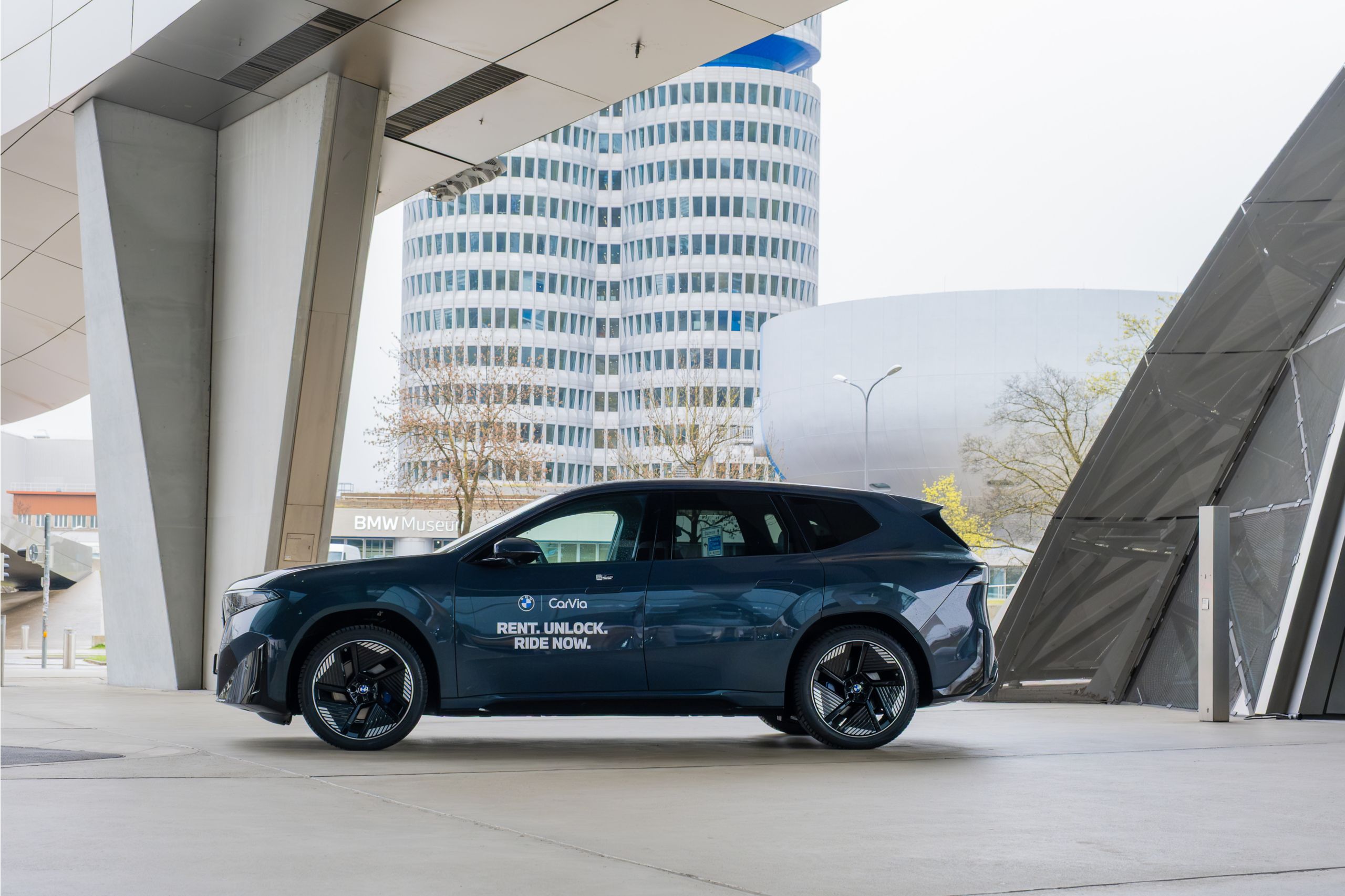 Dunkelblauer BMW iX3 mit Carsharing‑Branding steht unter moderner Architektur vor dem BMW Vierzylinder in München