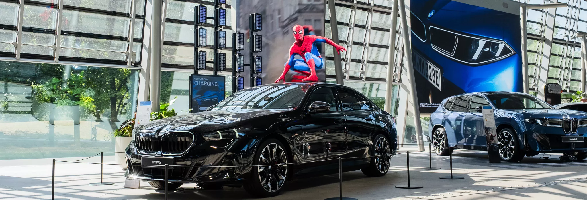 Spider-Man figure positioned on the roof of a BMW 5 at BMW Welt in front of a large digital Spider-Man display.