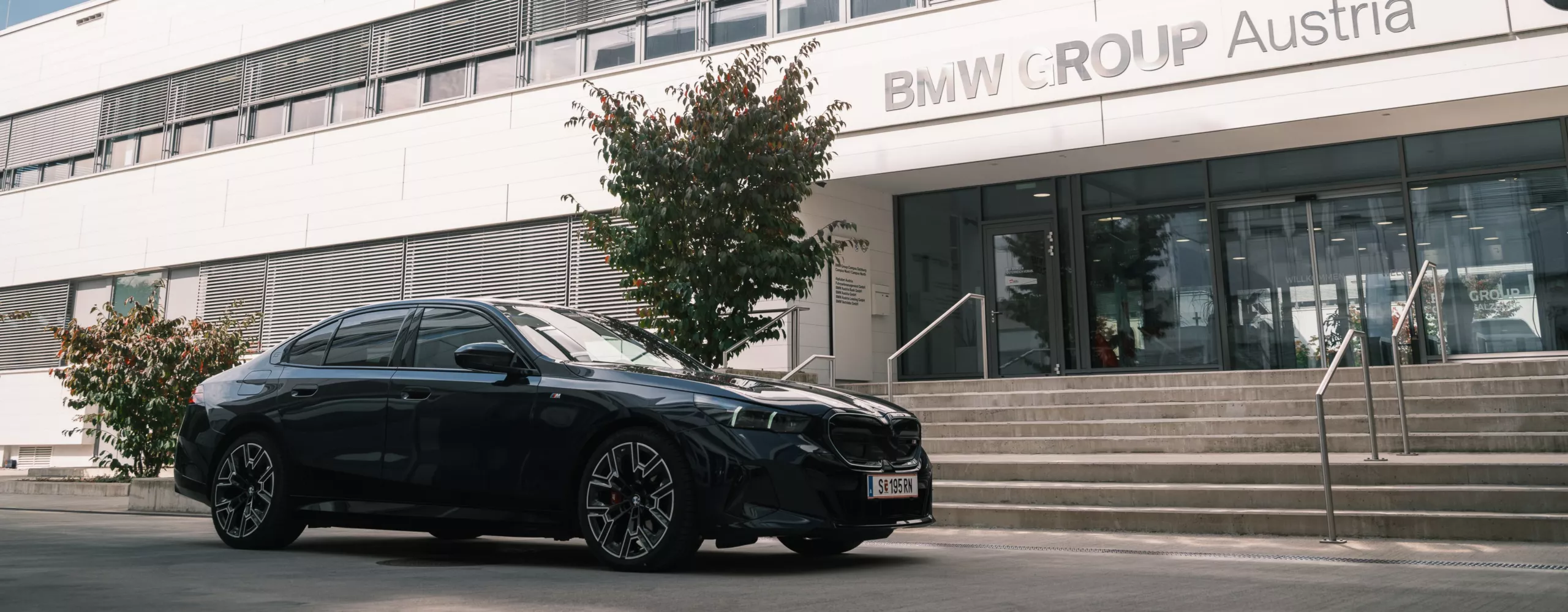 Ein dunkelbaluer BMW i5 steht vor den Treppen des Eingangs BMW Group Standort Salzburg Campus Nord. 