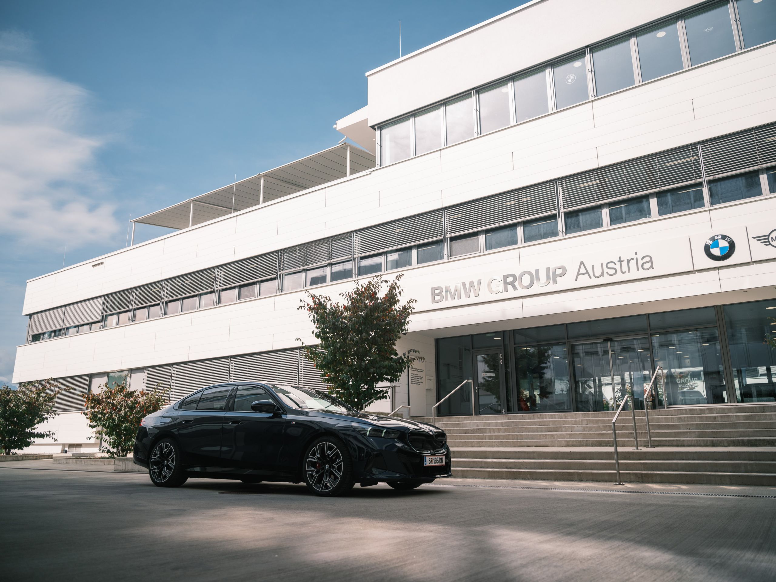 Ein dunkelbaluer BMW i5 steht vor den Treppen des Eingangs BMW Group Standort Salzburg Campus Nord. 