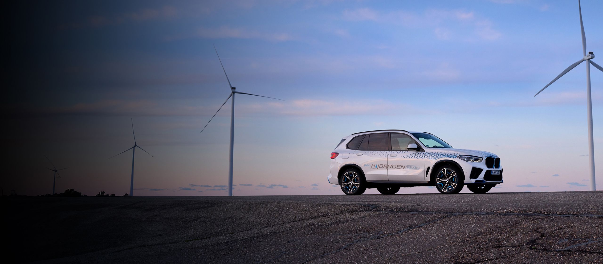 A BMW iX5 Hyrdogen Concept Vehicle is parked on an asphalt surface with wind turbines in the background